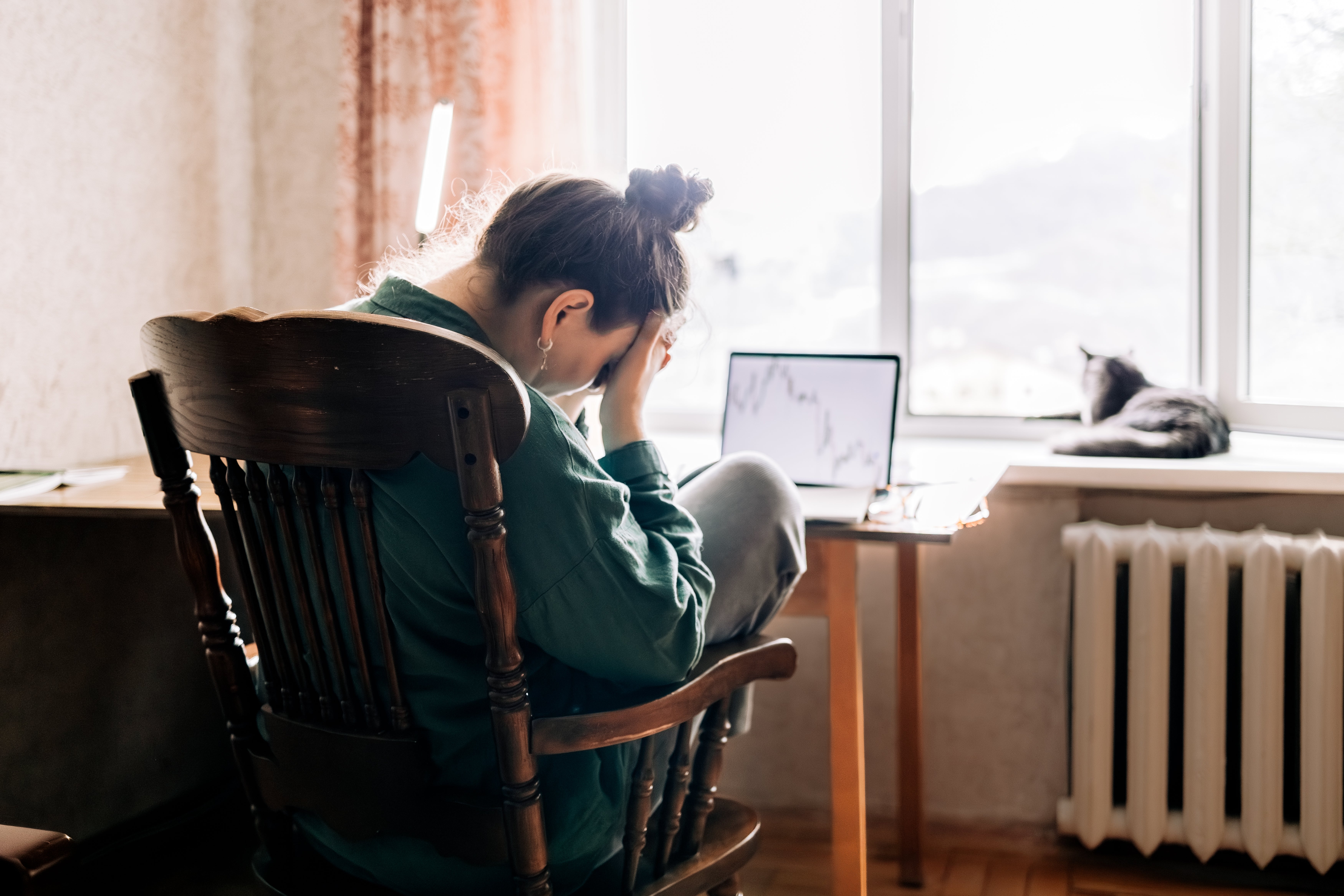 Person sitting successful  a chair, holding their caput  successful  their hands, with a laptop connected  a table  and a feline  connected  the windowsill successful  the background