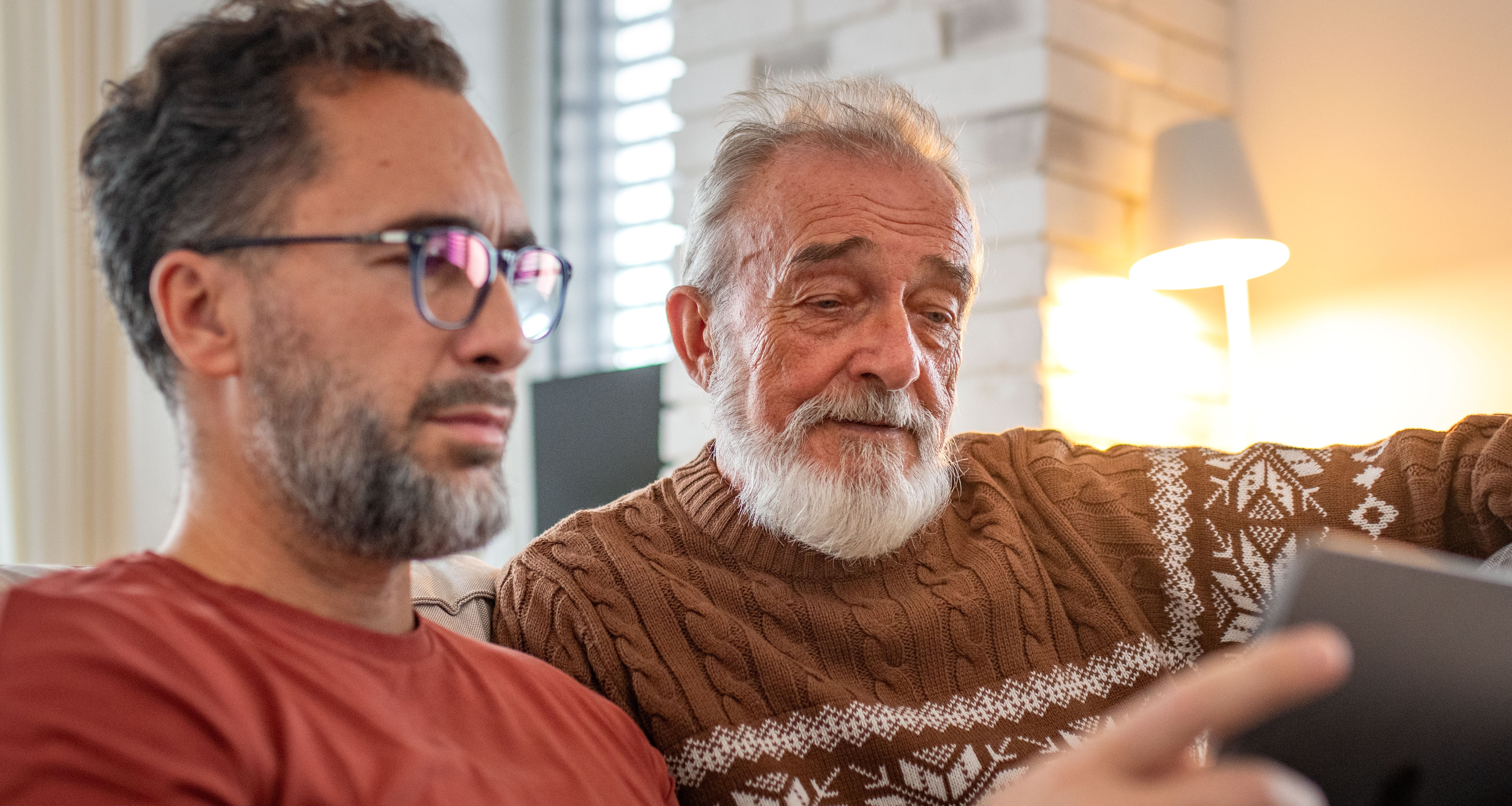 Two men sitting connected  a sofa  utilizing a tablet, 1  wearing glasses and a reddish  shirt, the different   successful  a knit sweater. They look  engaged with the screen