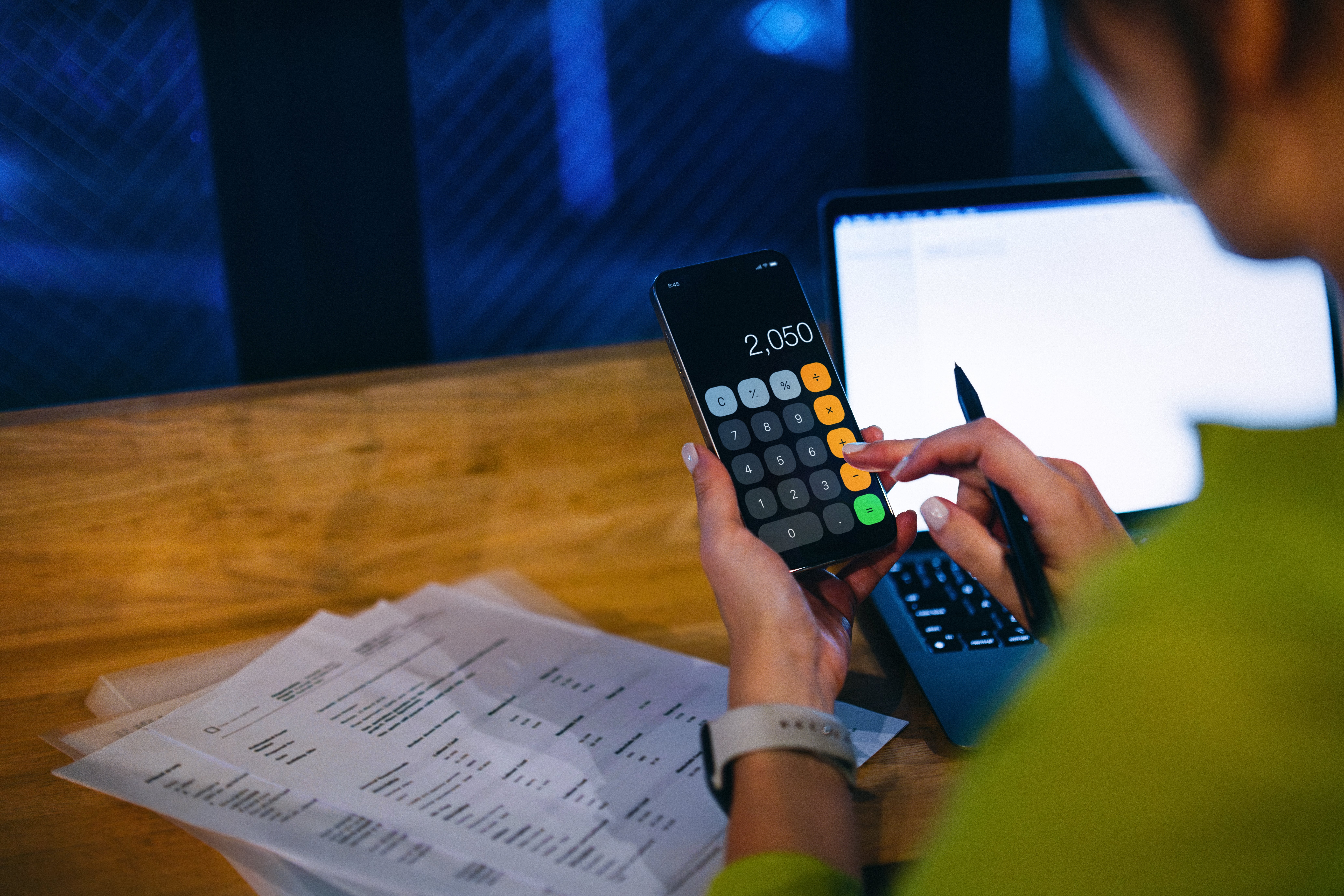 Person utilizing a smartphone calculator and laptop astatine  a woody  desk, surrounded by fiscal  documents