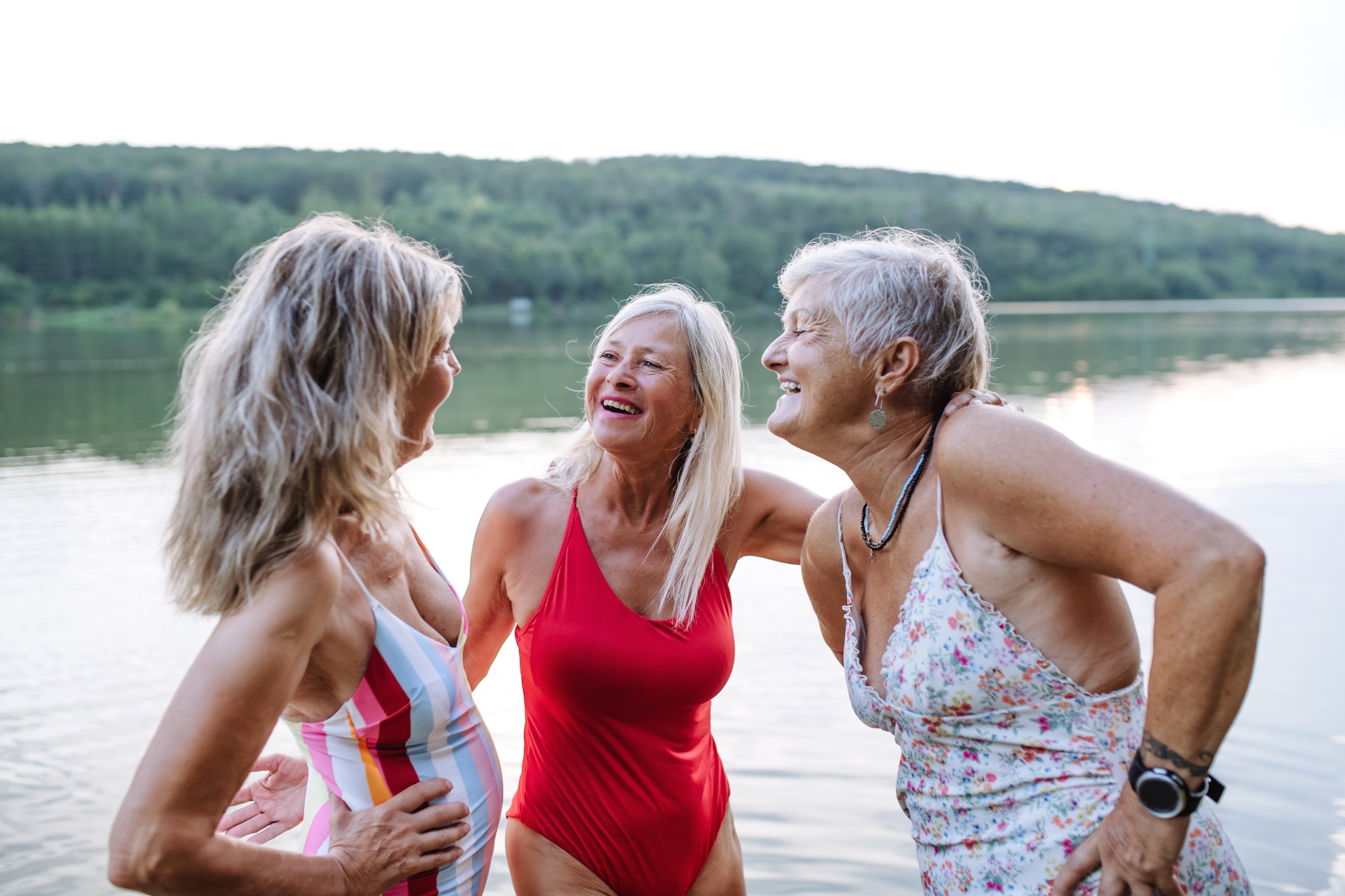 Three women successful  swimsuits grin  and chat by a lakeside, with trees successful  the background