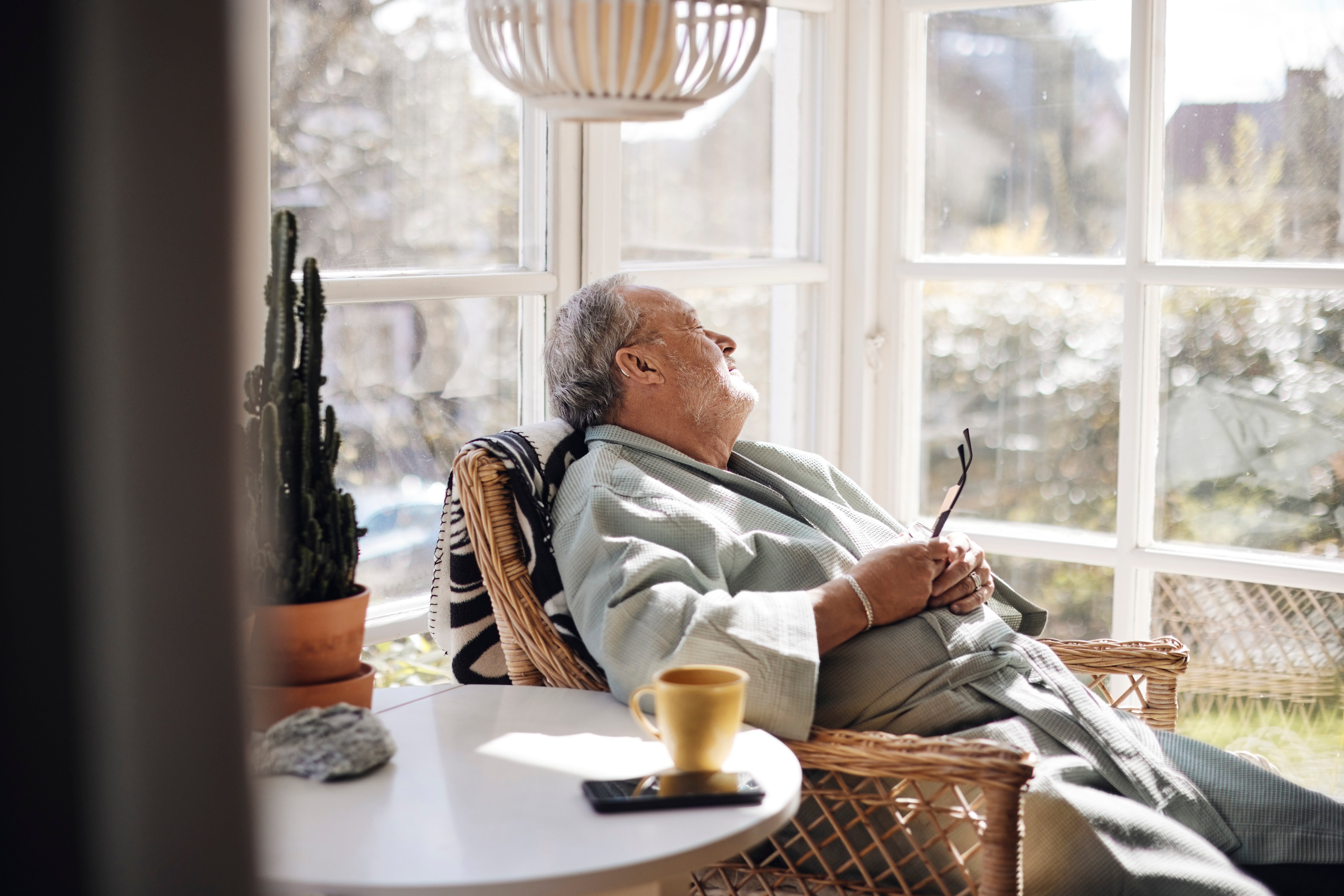An older antheral   successful  a robe relaxes successful  a sunlit seat  by a window, holding glasses. A cactus, table, and mug are nearby