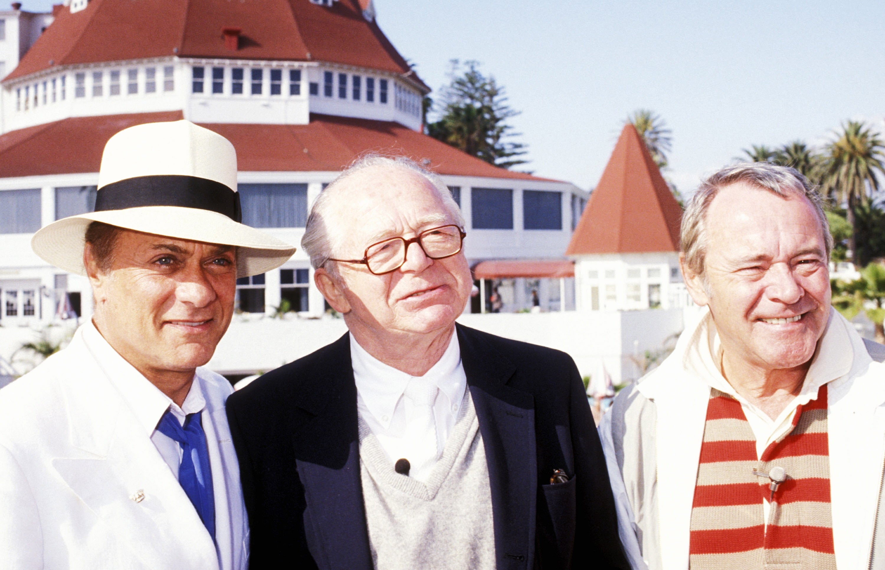 Three individuals basal   extracurricular  a ample  coastal building. One wears a achromatic  suit   and hat; 1  wears a overgarment  and glasses; 1  wears a striped garment  and blazer