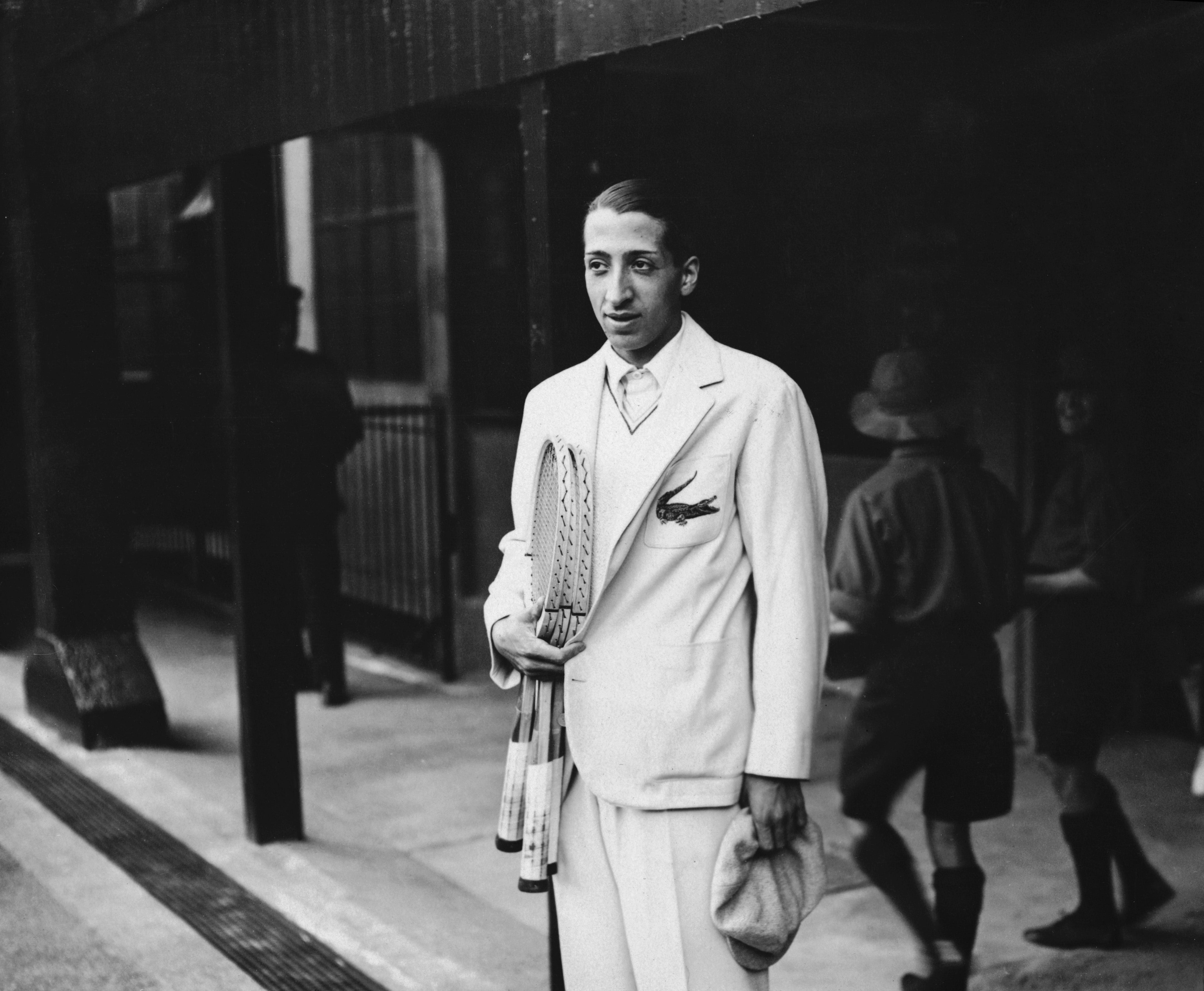 A antheral   successful  a vintage tennis outfit holds rackets, wearing a suit   with an emblem, lasting  extracurricular  a building. Retro style, aboriginal  sports context