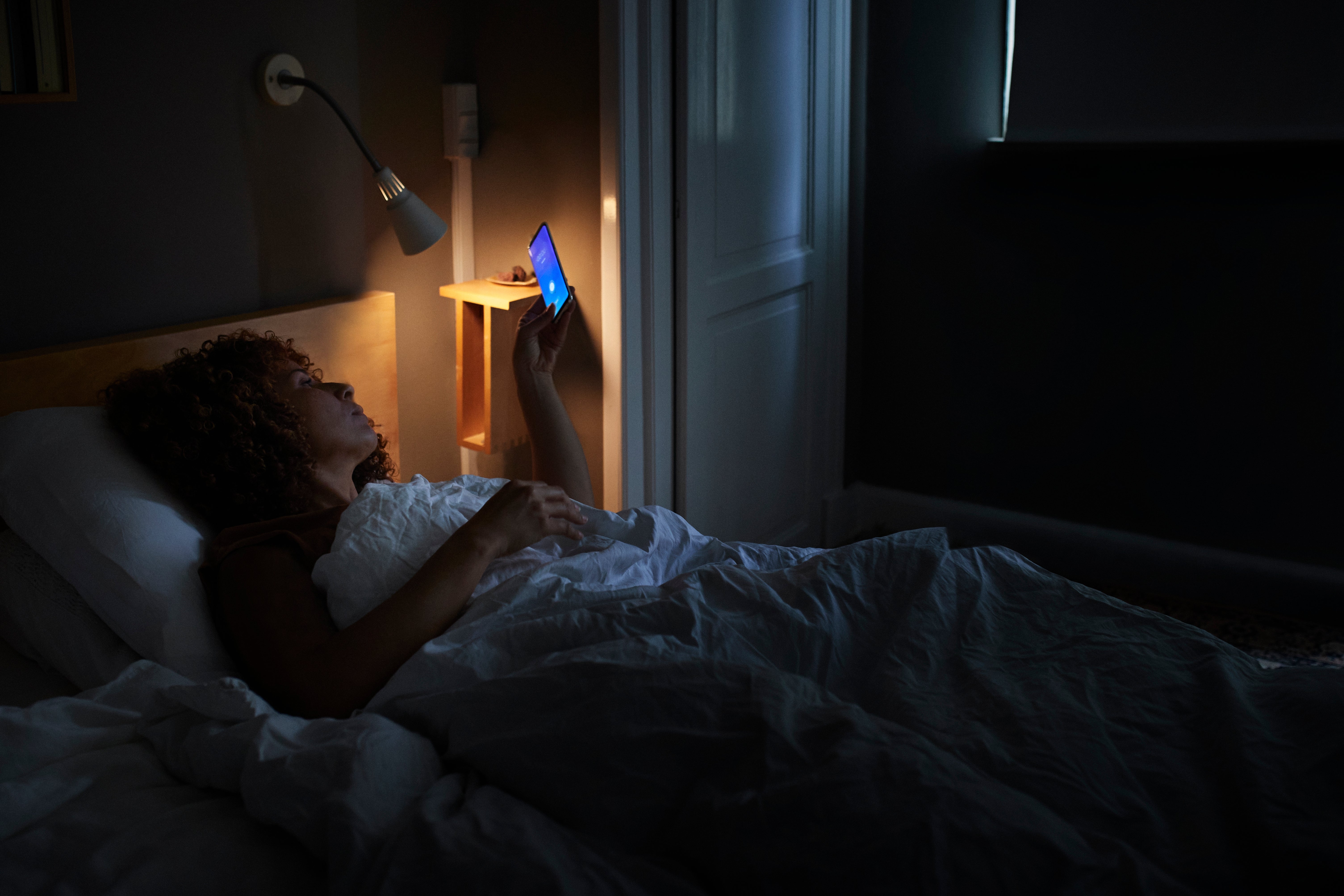 Person successful  bed, holding a telephone  with a focused expression. Room dimly lit by lamp, creating a relaxed nighttime atmosphere