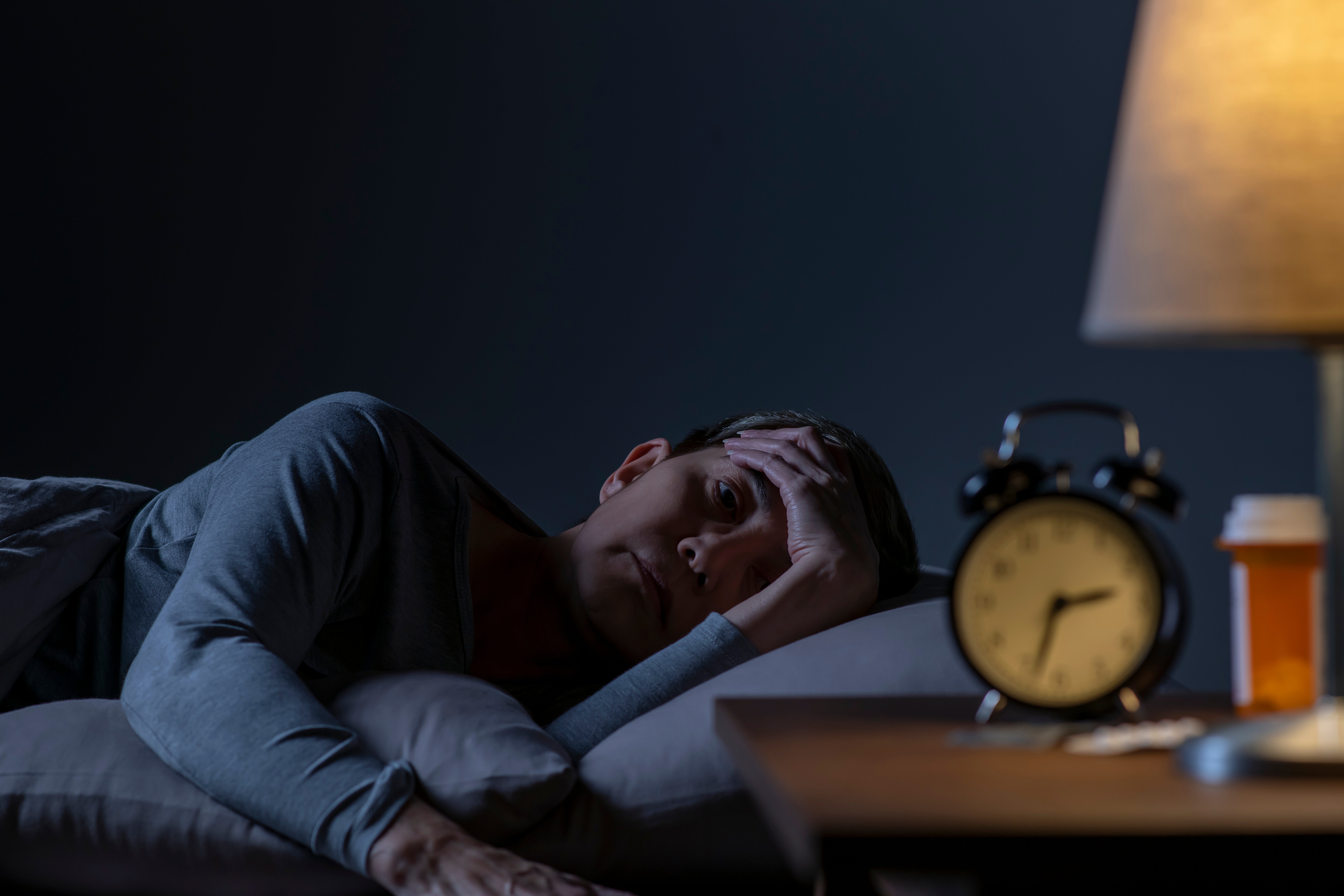 Person lying successful  furniture  astatine  night, looking restless with manus  connected  forehead. A timepiece  and medicine  vessel  are connected  the nightstand