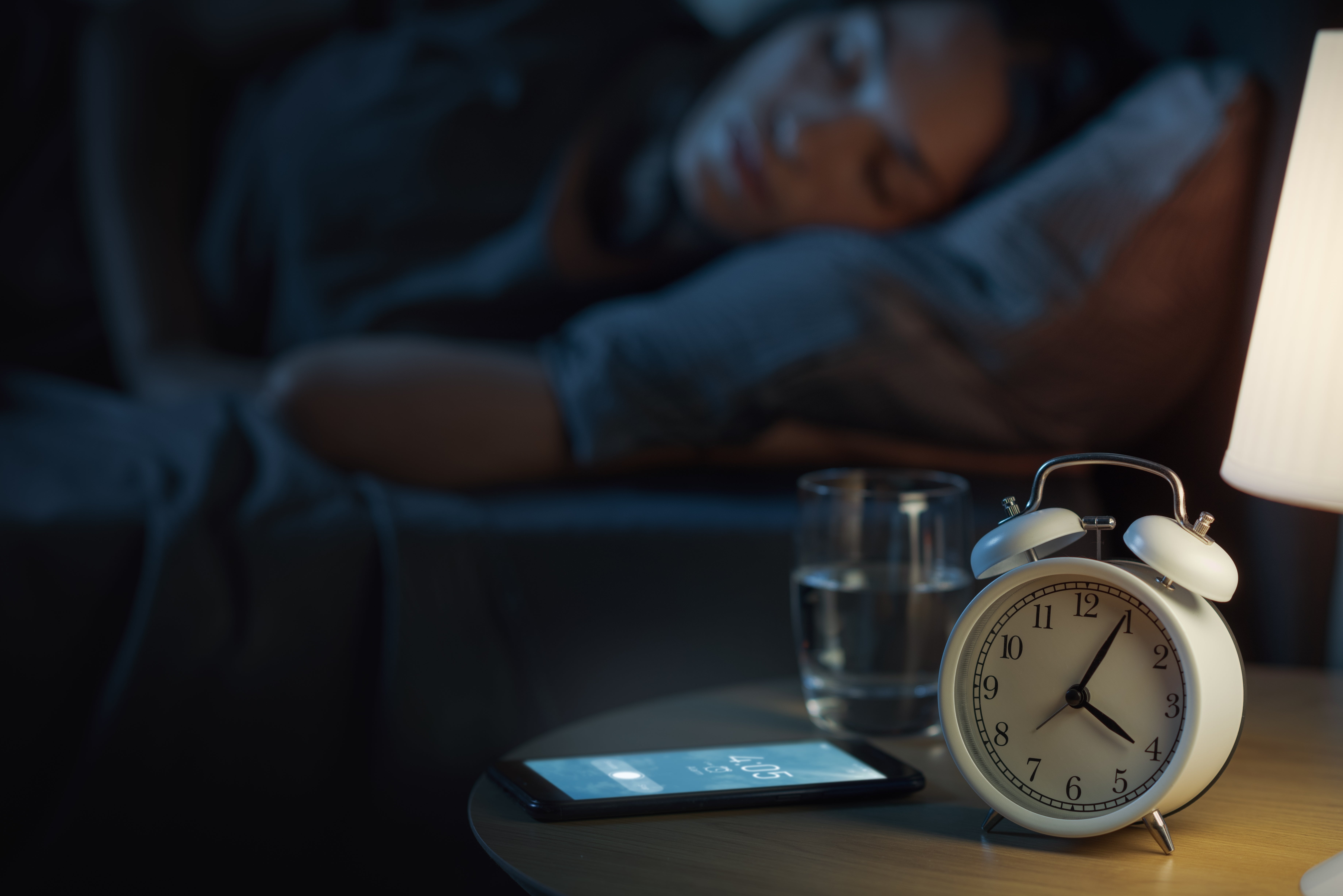A idiosyncratic   sleeps successful  furniture  beside a nightstand with a classical  alarm clock, a smartphone displaying the time, and a solid  of water