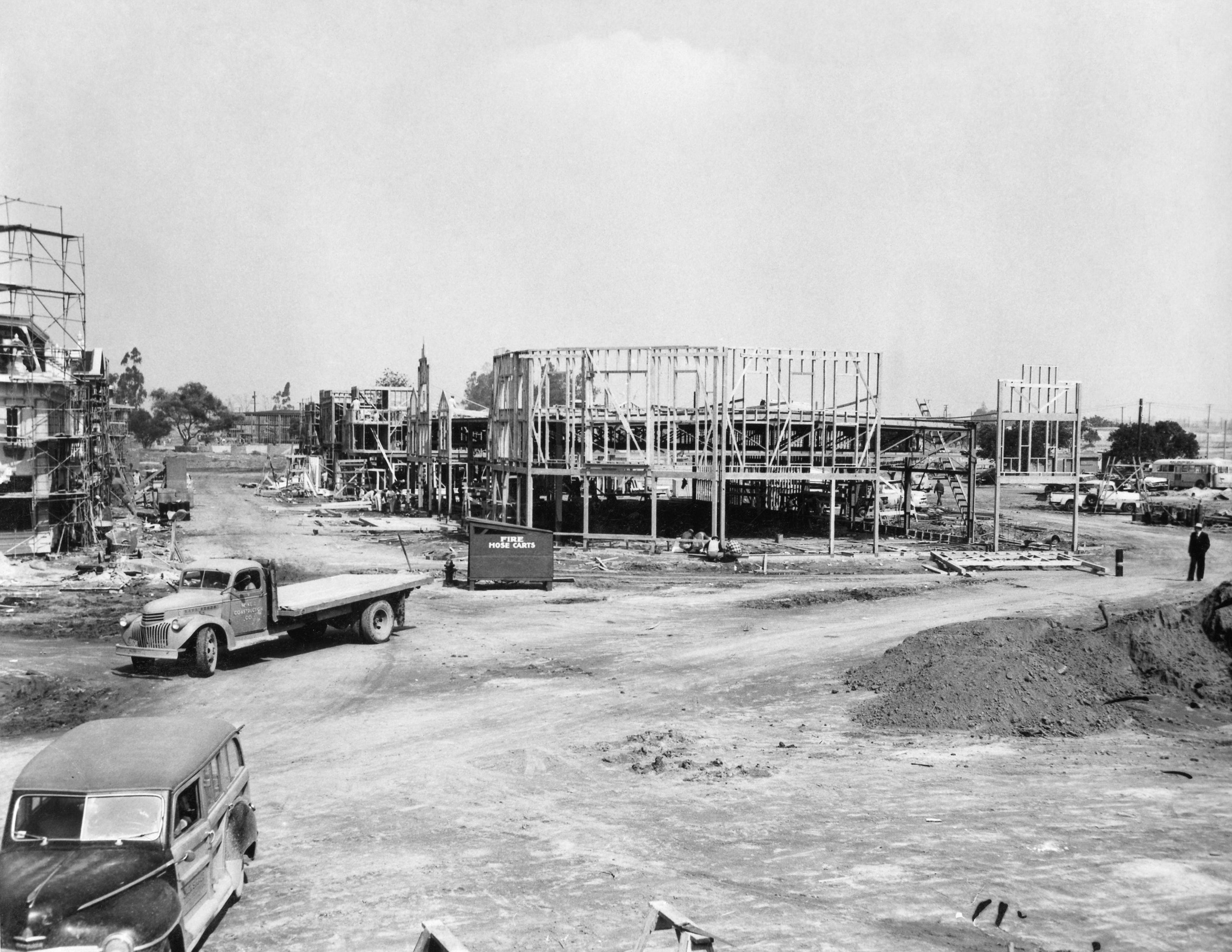 Construction tract  with woody  structures successful  progress, vintage cars, and scattered tools, suggesting an aboriginal  20th-century improvement  scene