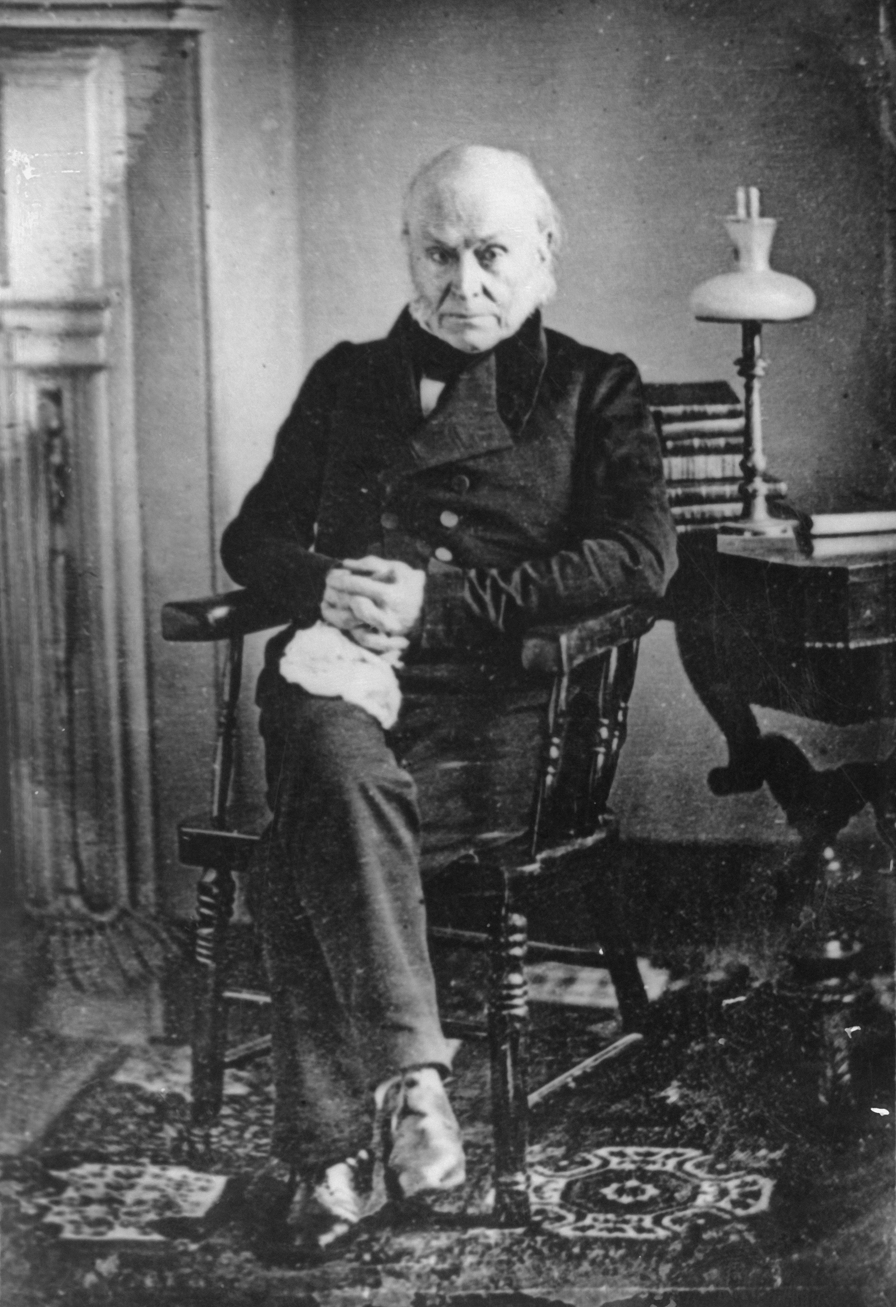 Older antheral   sitting successful  a woody  chair, wearing ceremonial  19th-century attire, with a stack of books and a lamp connected  a array  beside him