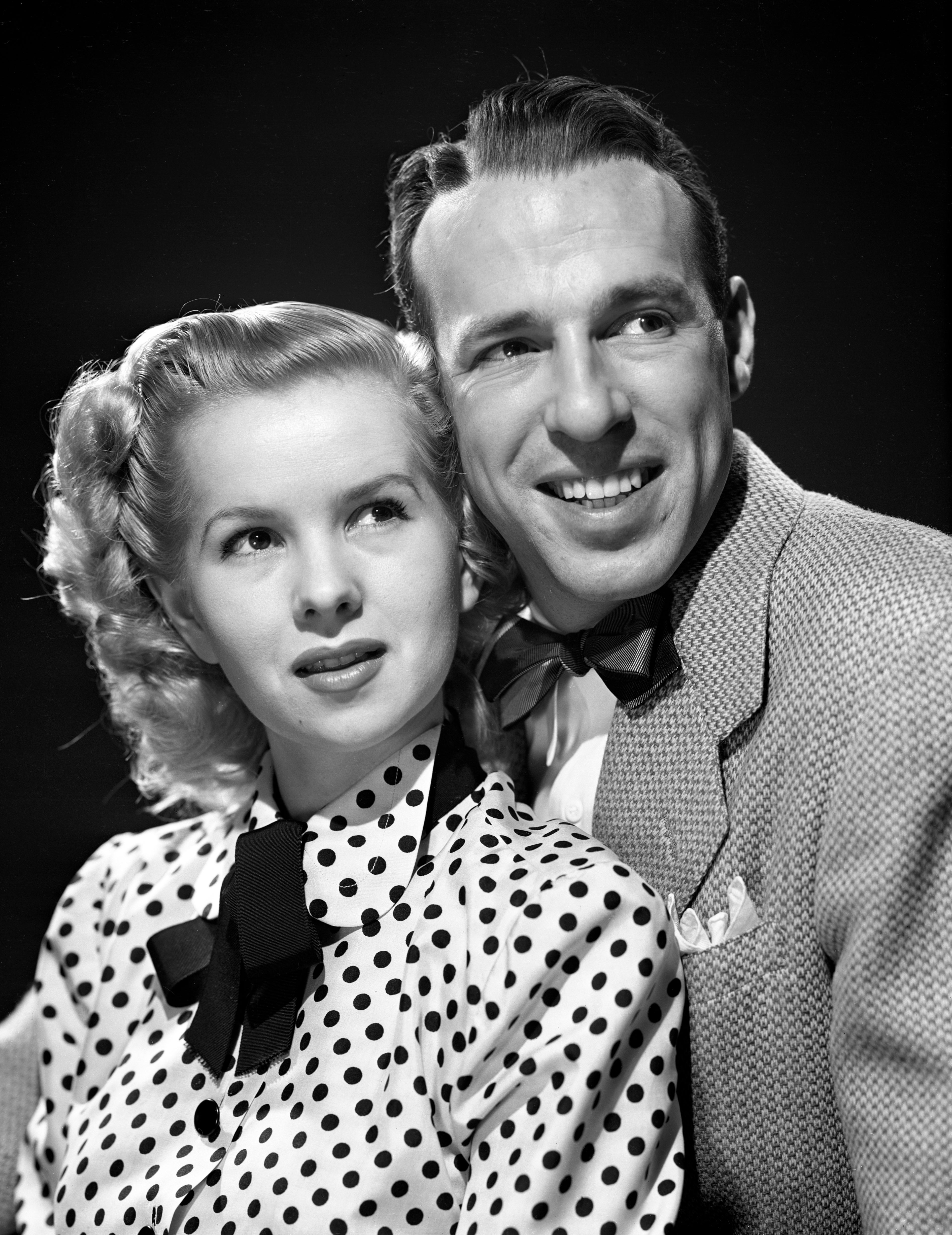 A vintage photograph  of a smiling couple, the pistillate   successful  a polka dot blouse with a bow, the antheral   successful  a suit   with a bow tie, looking off-camera
