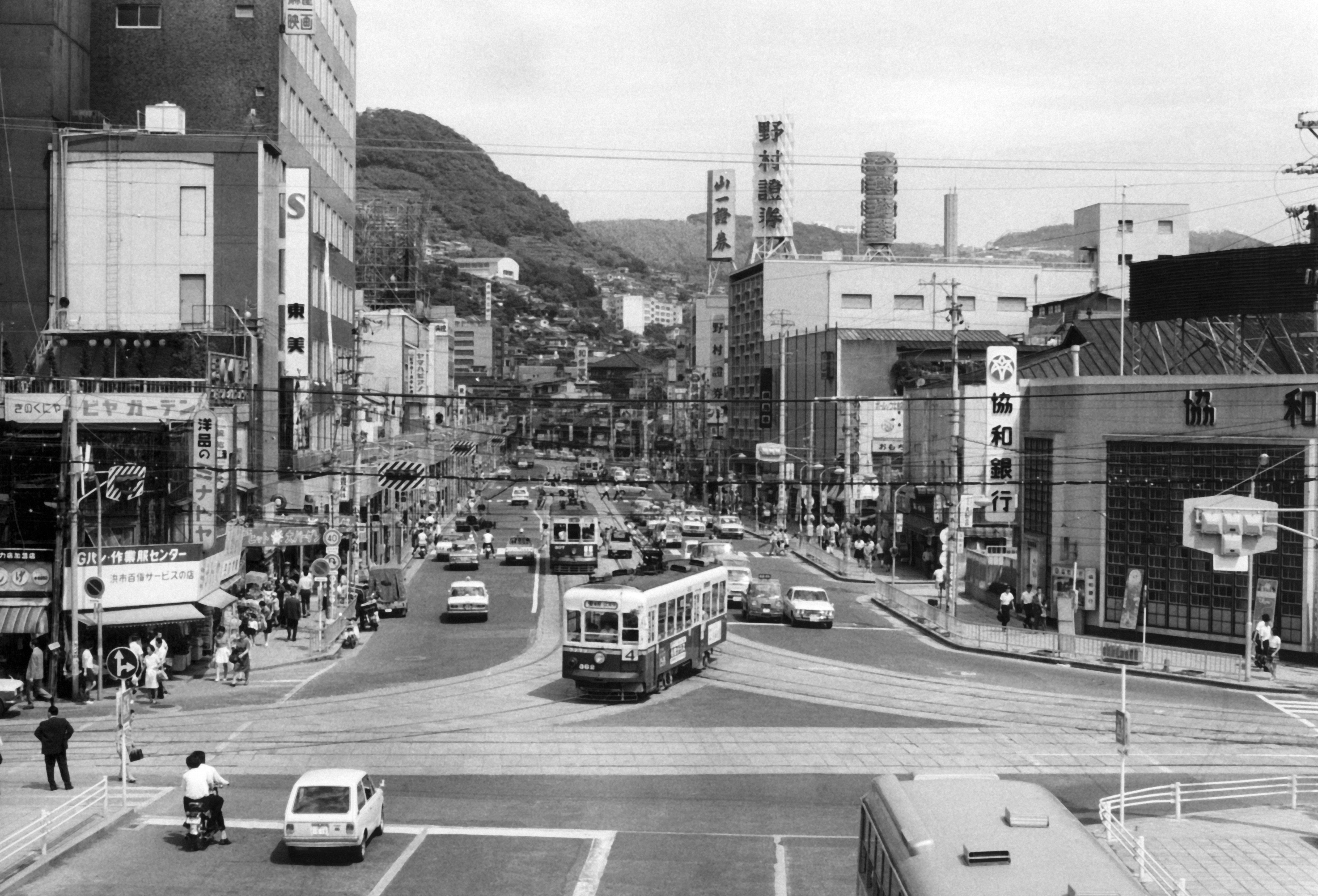 Busy 1960s Japanese thoroughfare  country   with cars, buses, and pedestrians, framed by municipality  buildings and distant   hills