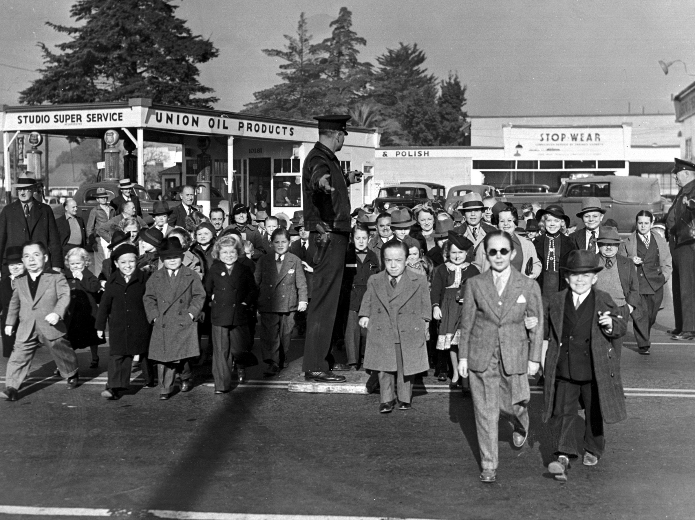 Historical photograph  of children crossing a thoroughfare  with a constabulary  serviceman  guiding them. Businesses successful  the inheritance  see  a work  presumption    and shops