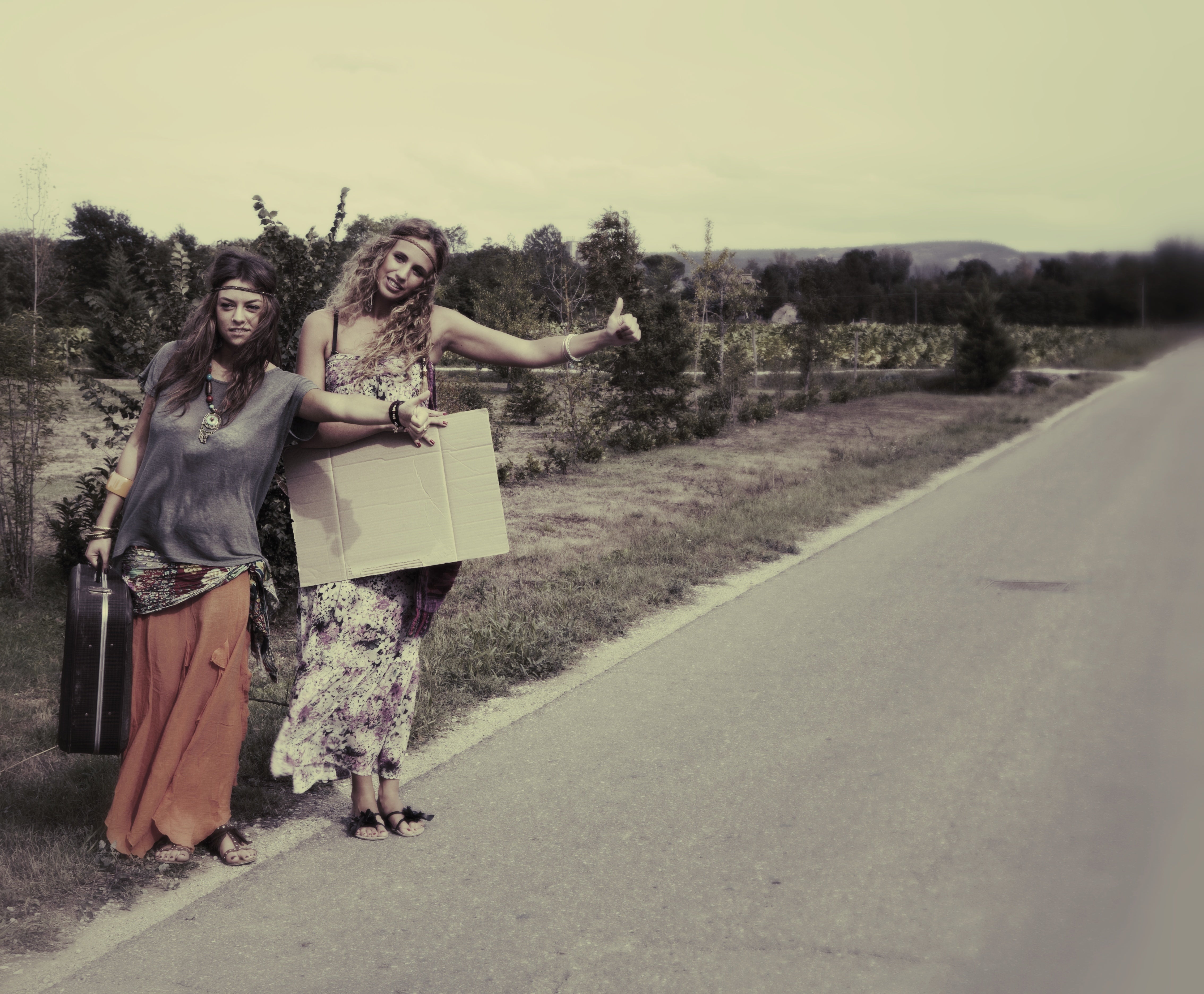 Two women successful  bohemian-style covering  hitchhiking connected  a agrarian  road, 1  holding a suitcase and a blank cardboard sign