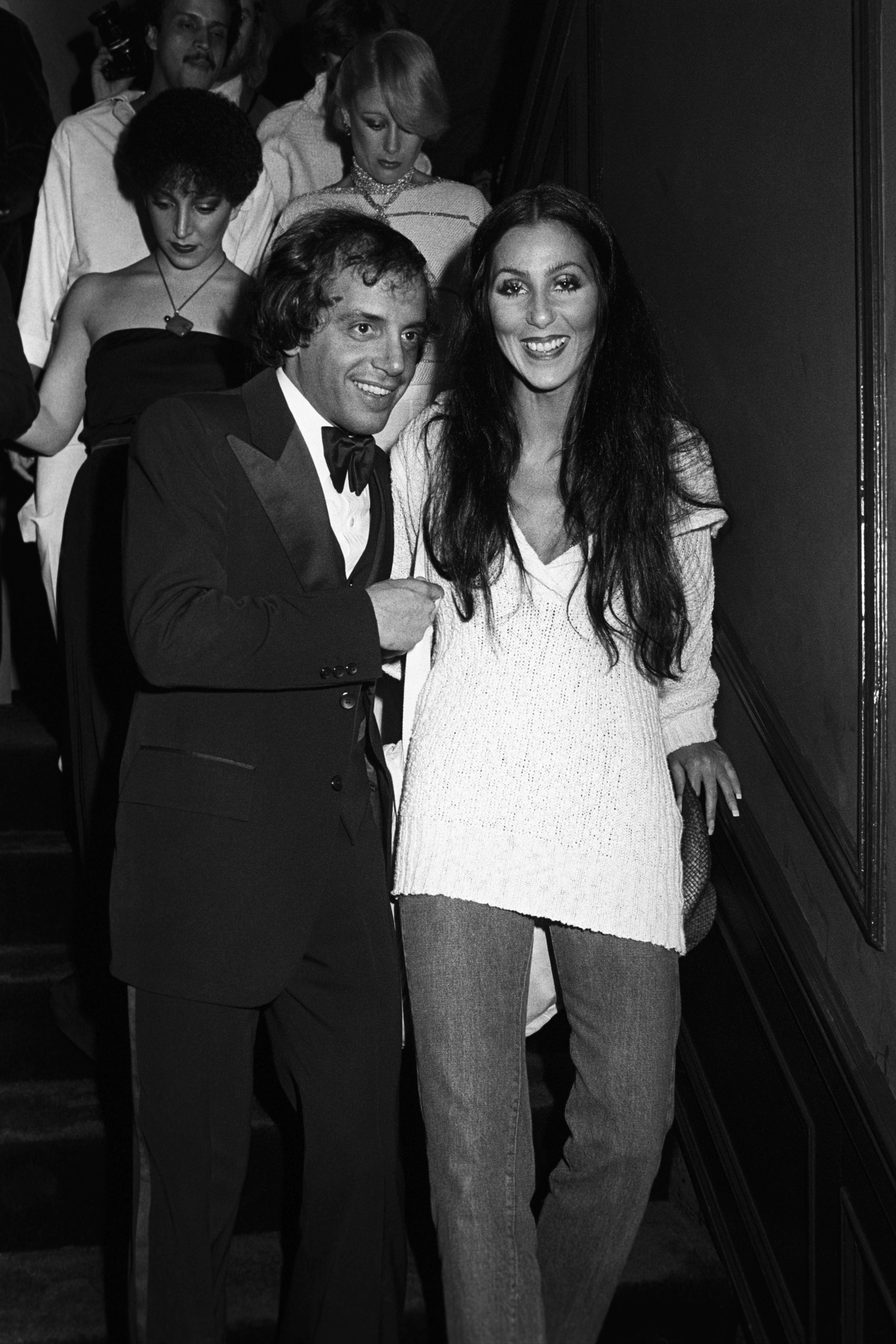 A pistillate   successful  casual attire with a sweater and pants posing adjacent  to a antheral   successful  a tuxedo, some  smiling, astatine  an lawsuit   with respective  radical   successful  the background