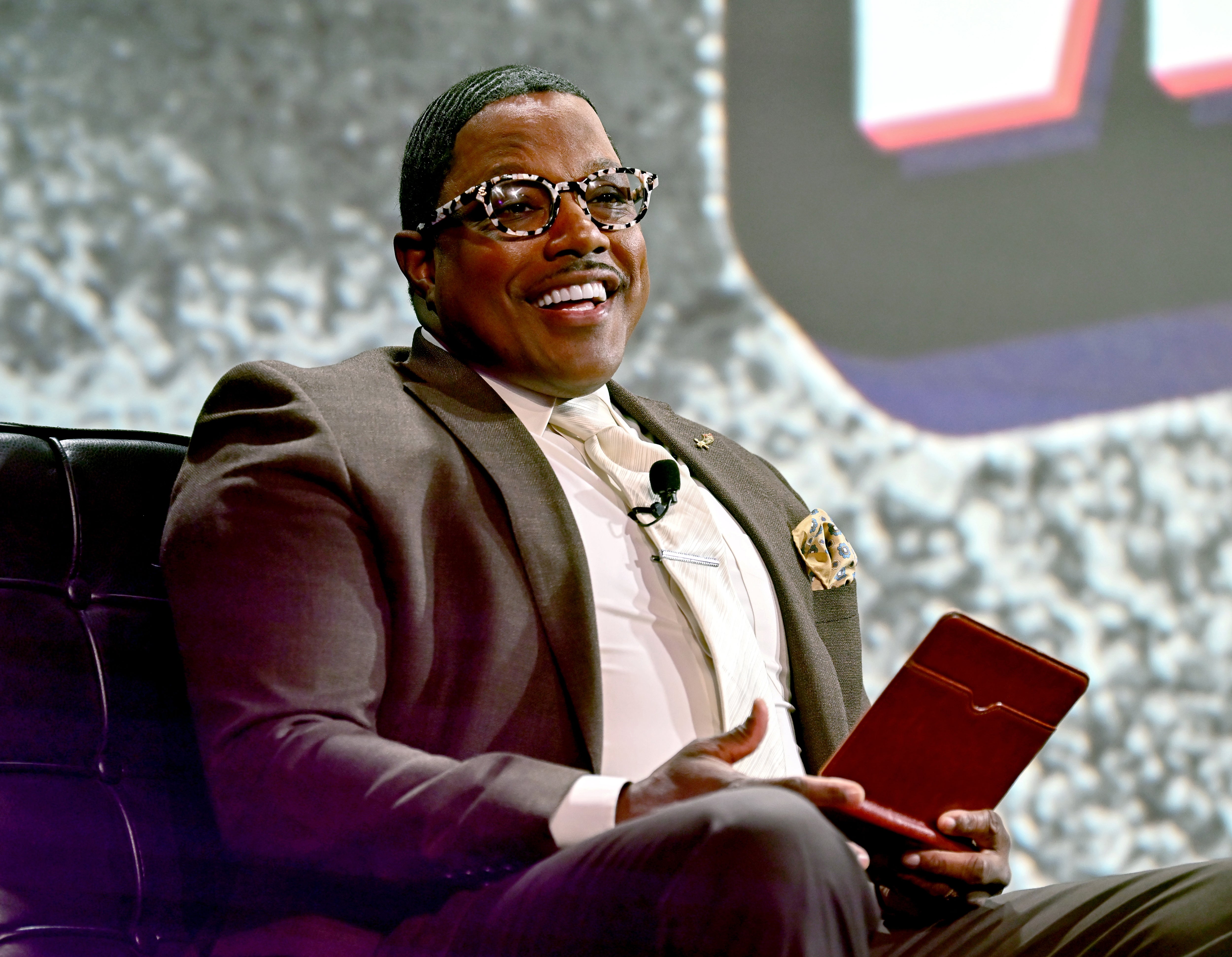 Smiling idiosyncratic   successful  a tailored suit   and patterned glasses sits holding a notebook, apt  astatine  a personage  lawsuit   oregon  interview