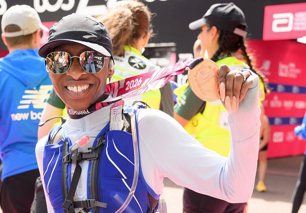Person proudly displaying a marathon medal, wearing a running vest and reflective sunglasses at a post-race event with other participants nearby