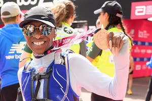 Person proudly displaying a marathon medal, wearing a running vest and reflective sunglasses at a post-race event with other participants nearby