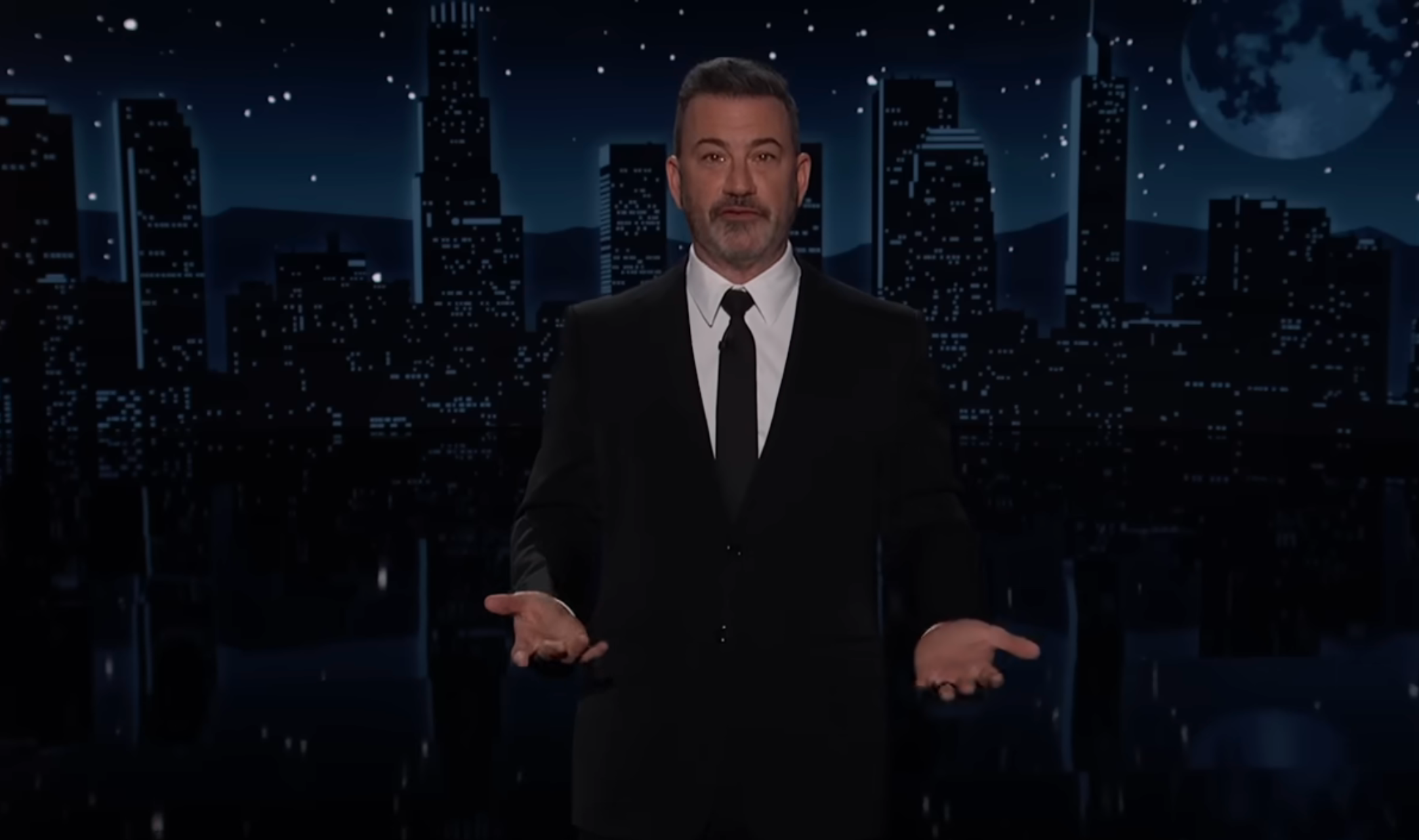 A antheral   successful  a suit   stands connected  a signifier    with a cityscape backdrop astatine  night, gesturing with his hands open