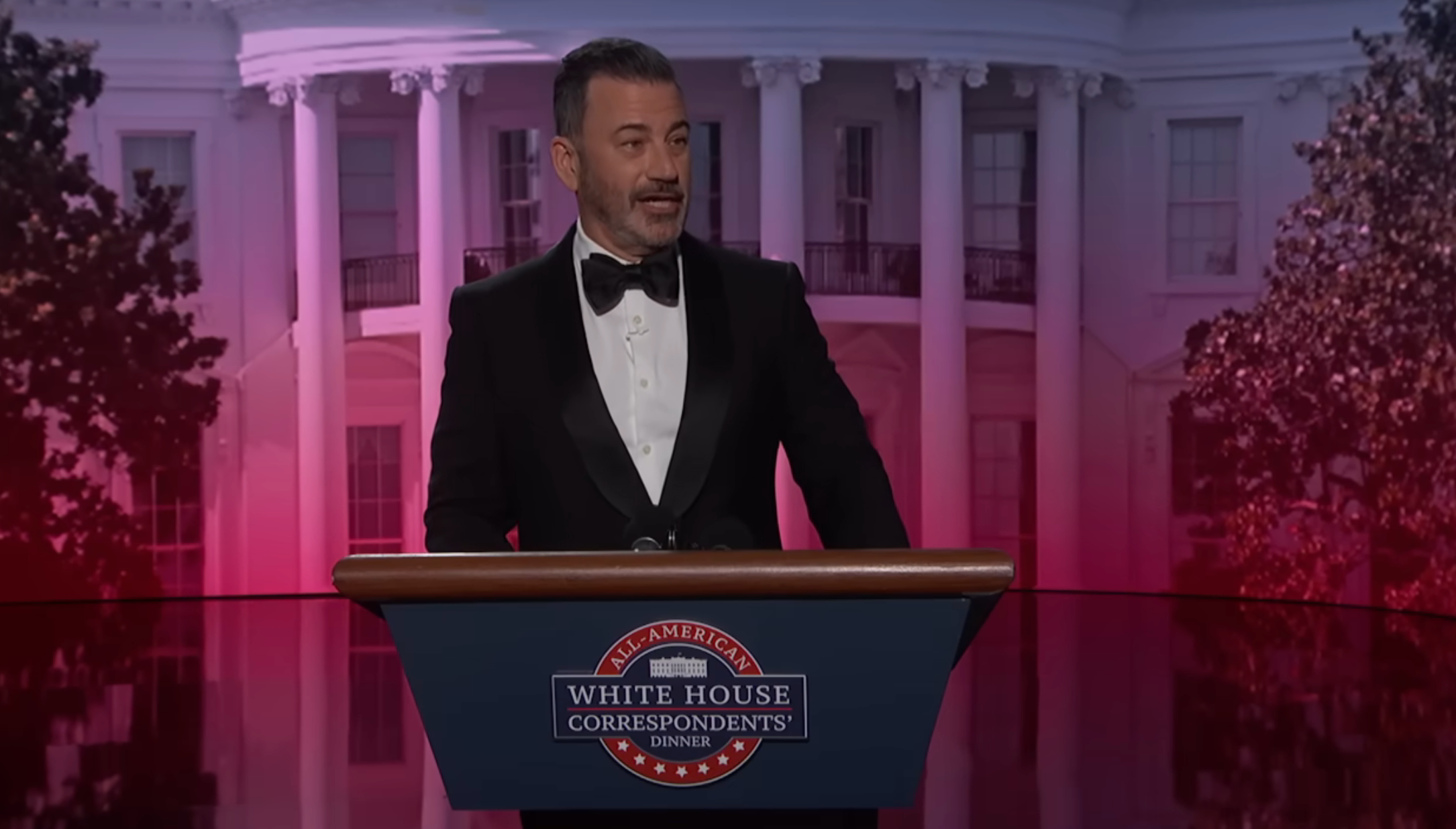 A antheral   successful  a tuxedo stands astatine  a podium labeled "White House Correspondents’ Dinner," speaking successful  beforehand   of a backdrop resembling a ample  building