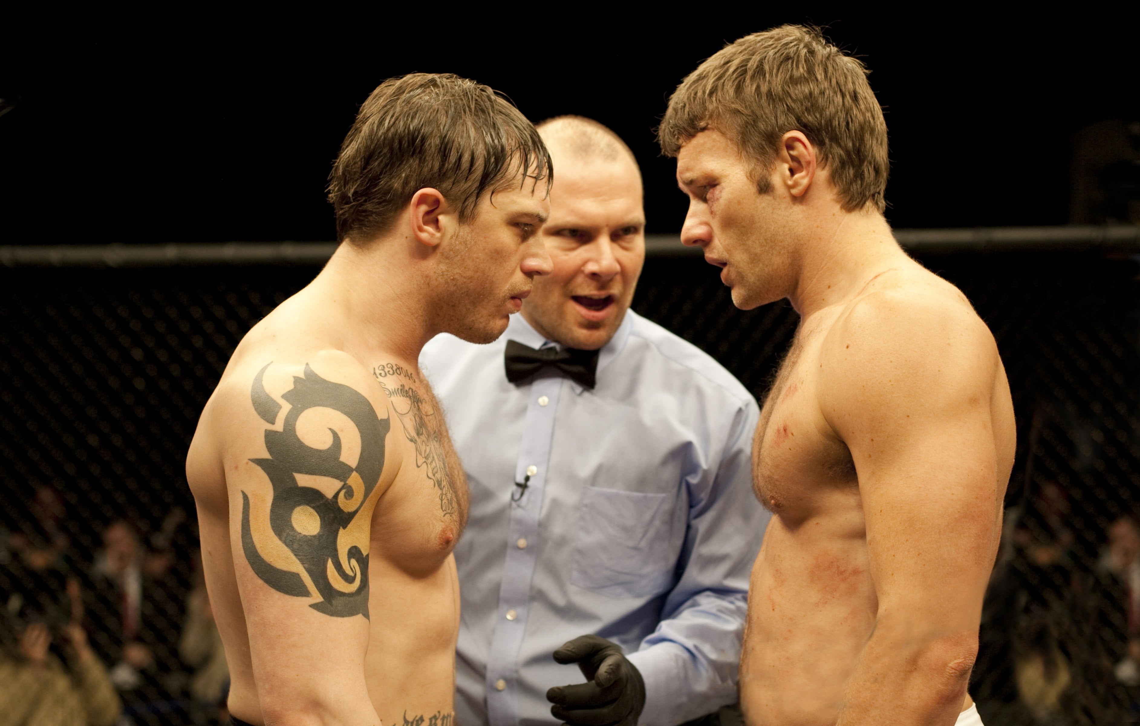 Two male fighters, shirtless, face off in a ring with a referee between them, suggesting a tense, competitive moment in a fighting event