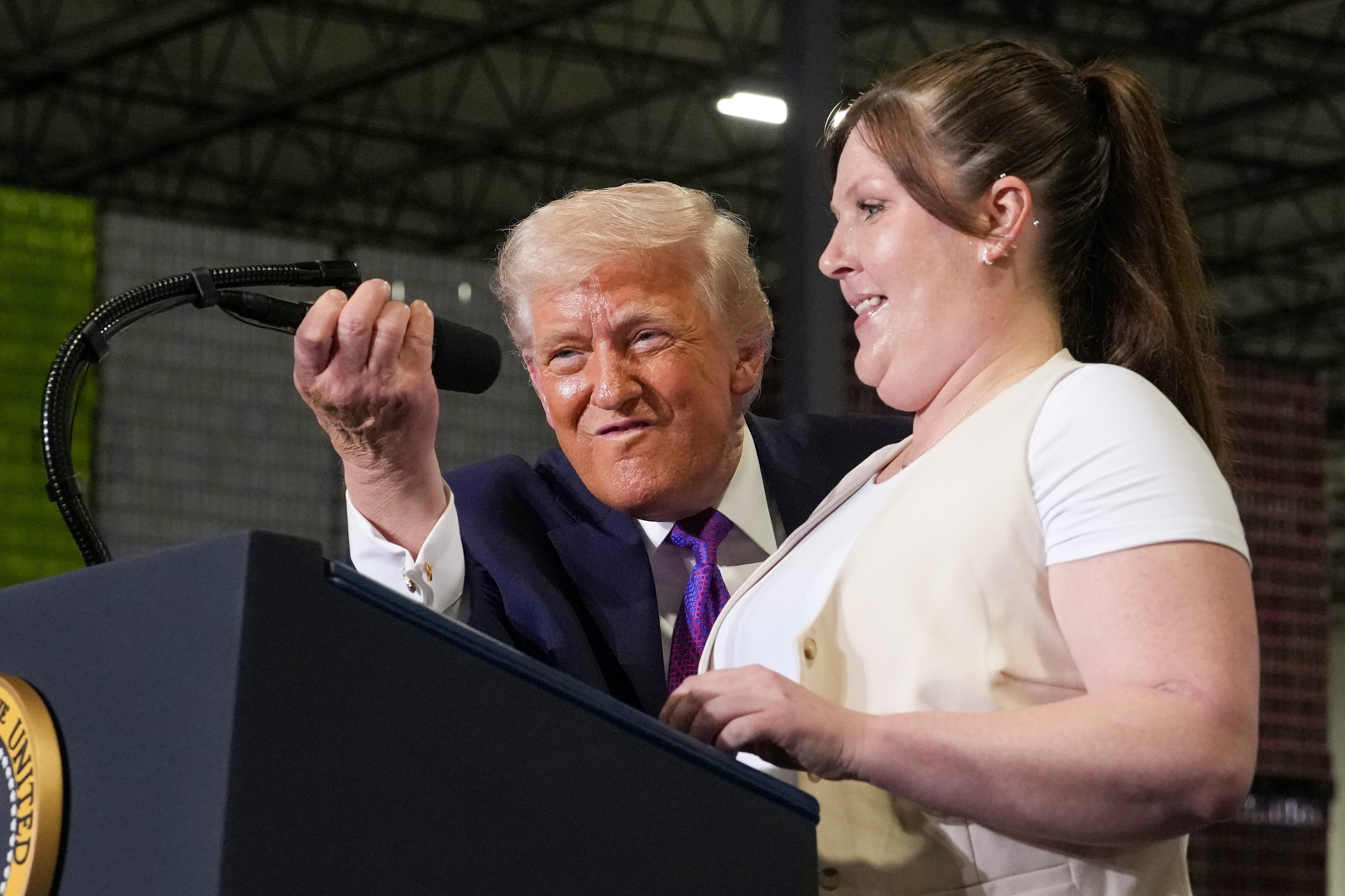 Donald Trump justerer en mikrofon med en smilende kvinne på et innendørs arrangement, med et podium foran seg