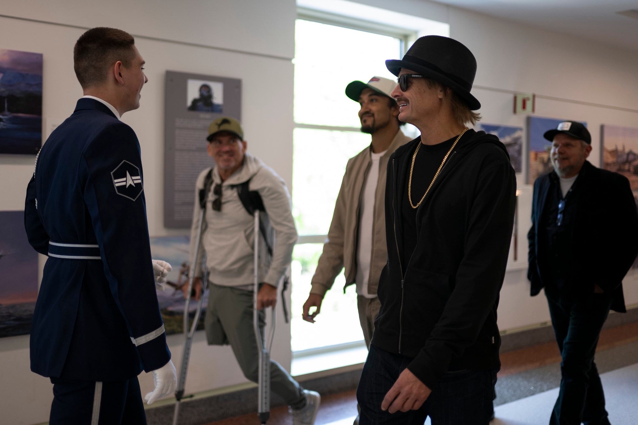 En gruppe mennesker, inkludert en mann på krykker og en mann i dress, går i en solfylt gang og snakker med en uniformert person