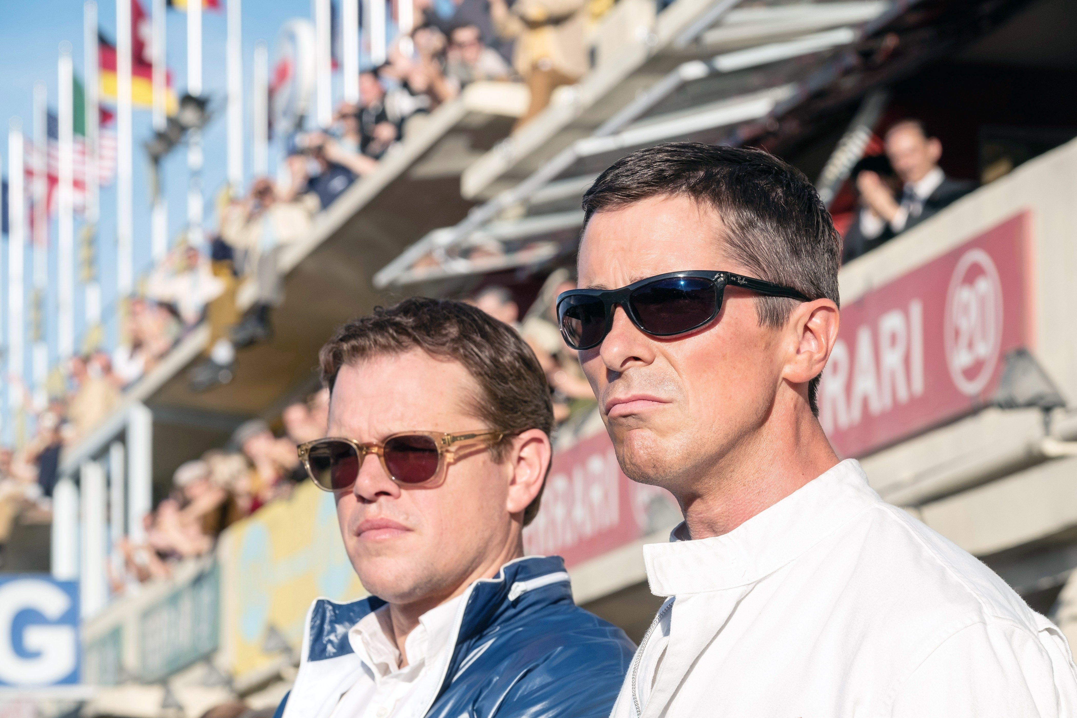 Two men in sunglasses stand at a racing event, focused on the track. The image captures a moment of intense concentration