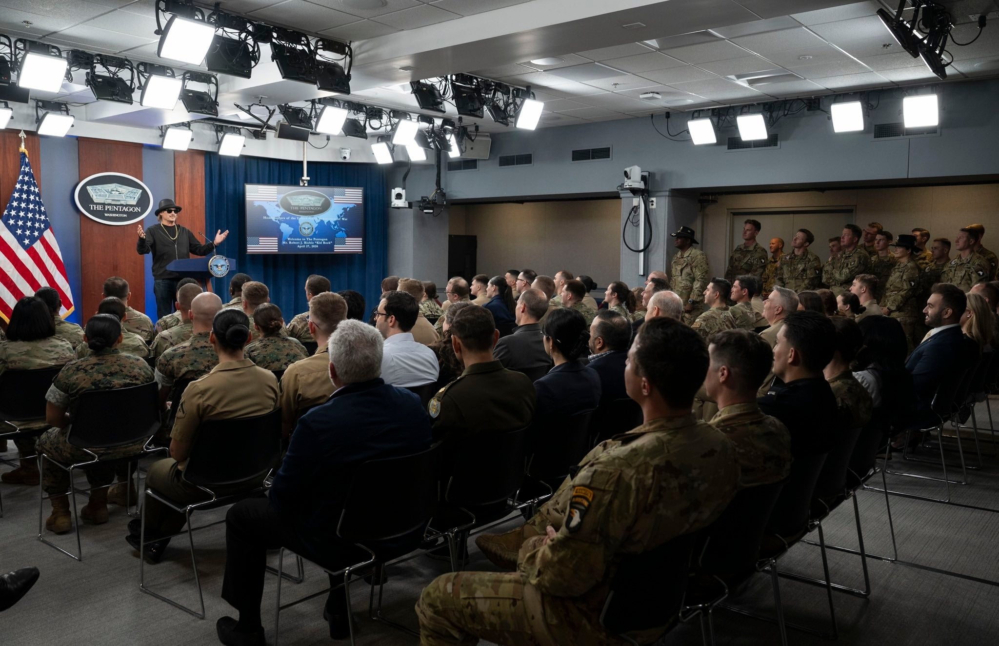 En militær briefing finner sted i et konferanserom med uniformert personell, en foredragsholder ved et podium og en presentasjonsskjerm
