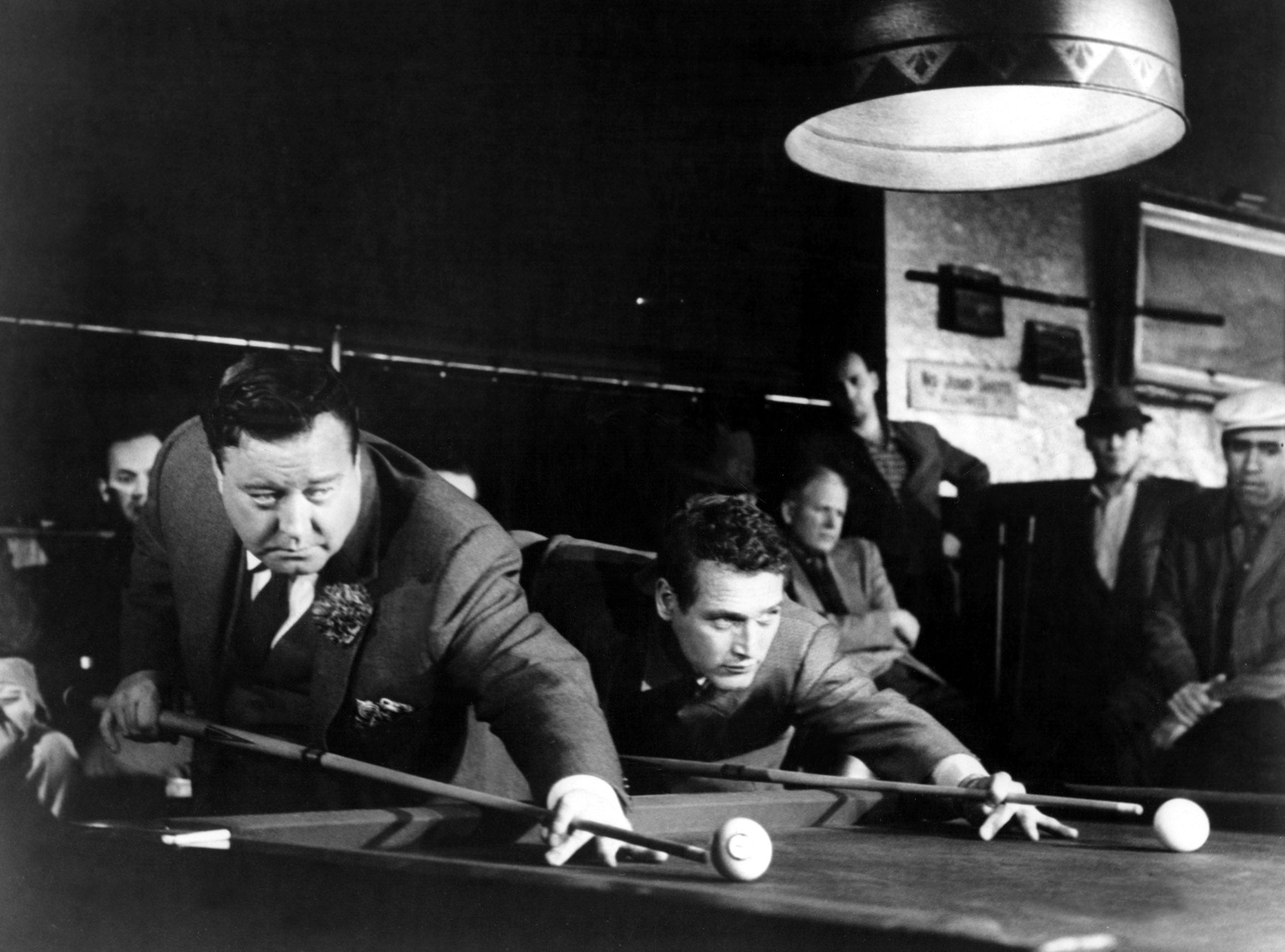 Two actors focus intensely while playing pool in a dramatic scene, surrounded by onlookers in a dimly lit space