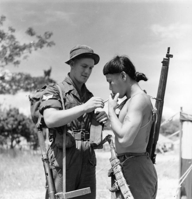 Two soldiers, 1  successful  a subject   azygous   and the different   shirtless, are lasting  outdoors. The uniformed worker  is lighting a cigaret  for the other