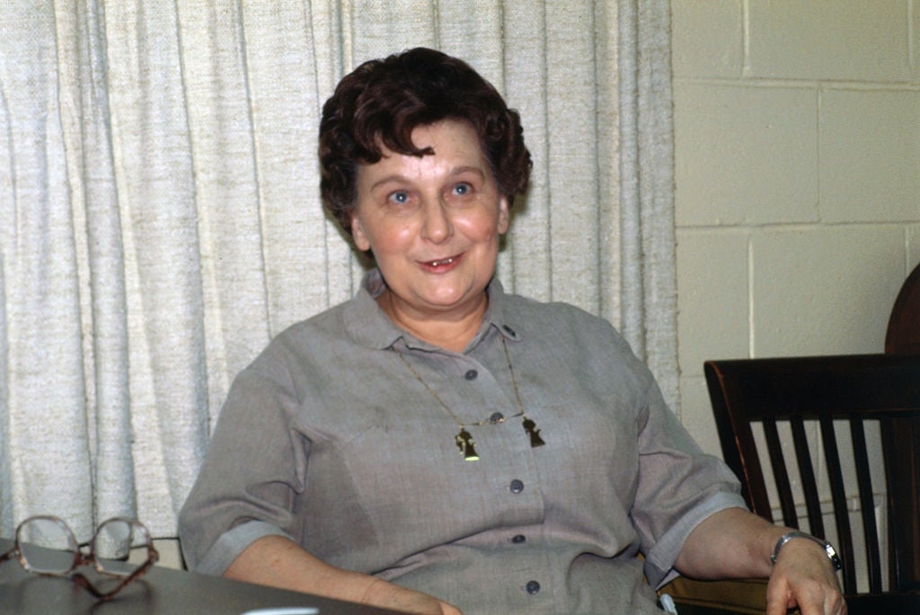 Person seated indoors, smiling, wearing a button-up garment  and a necklace. Glasses are connected  the array  nearby