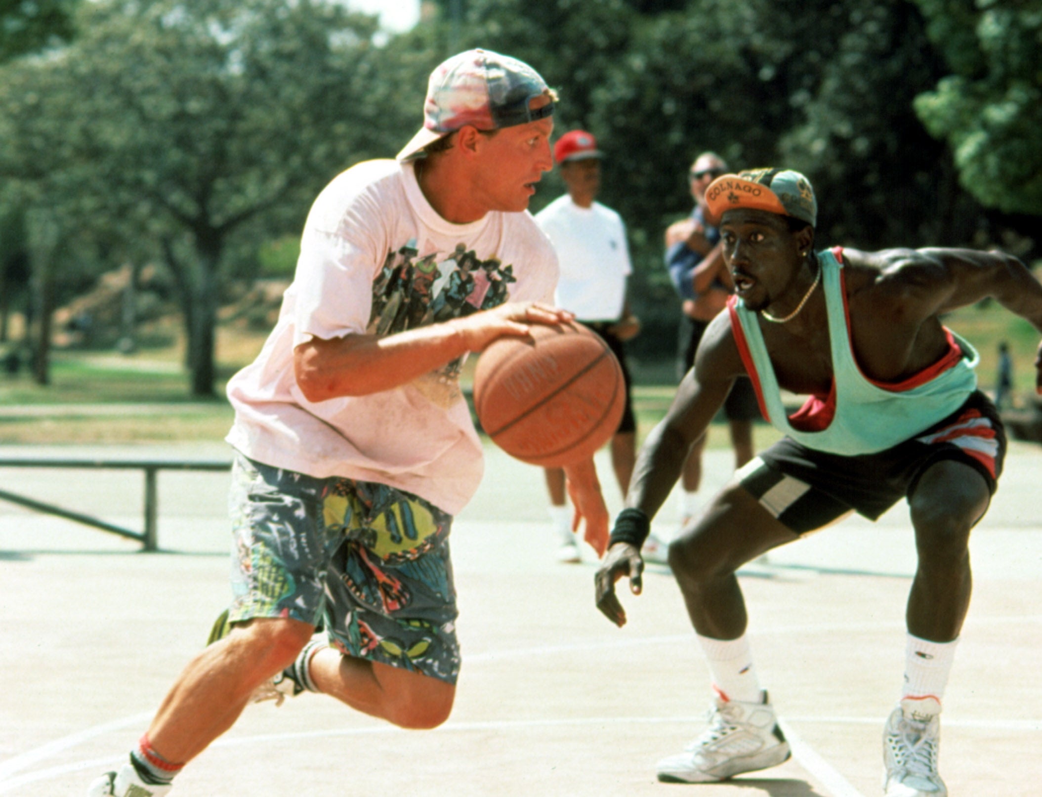 Two men playing basketball outdoors; one is dribbling while the other defends him. Casual street style clothing suggests a laid-back game