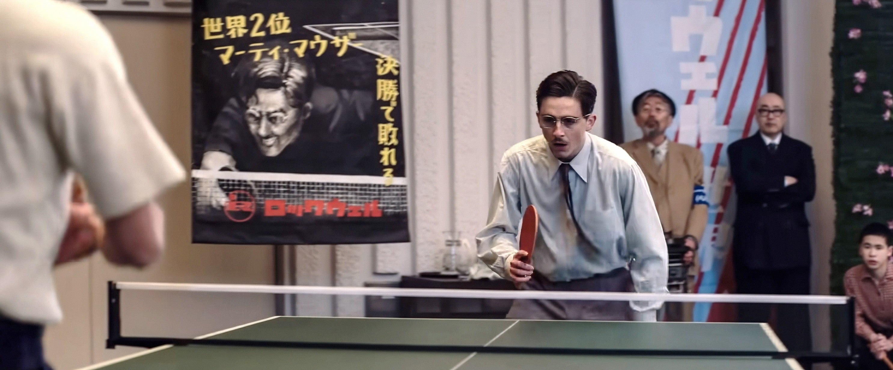 A man in vintage attire playing table tennis indoors, with a tournament banner and spectators in the background