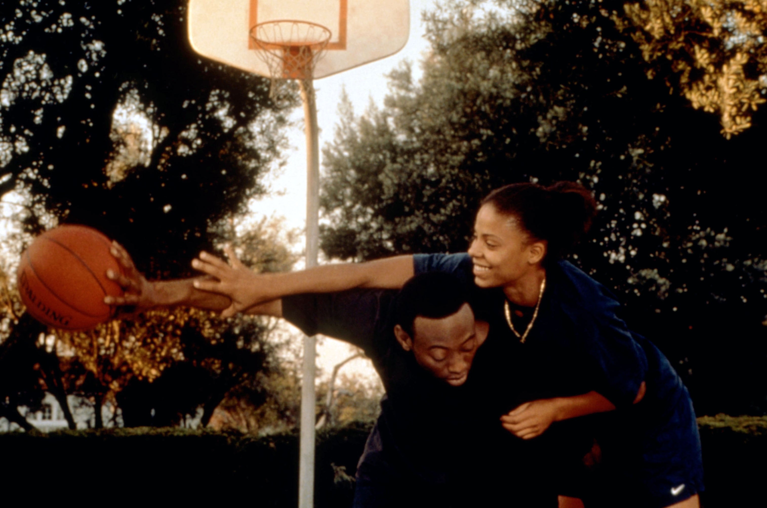 Two people playing basketball outdoors, one trying to block a pass. Trees and a basketball hoop are in the background