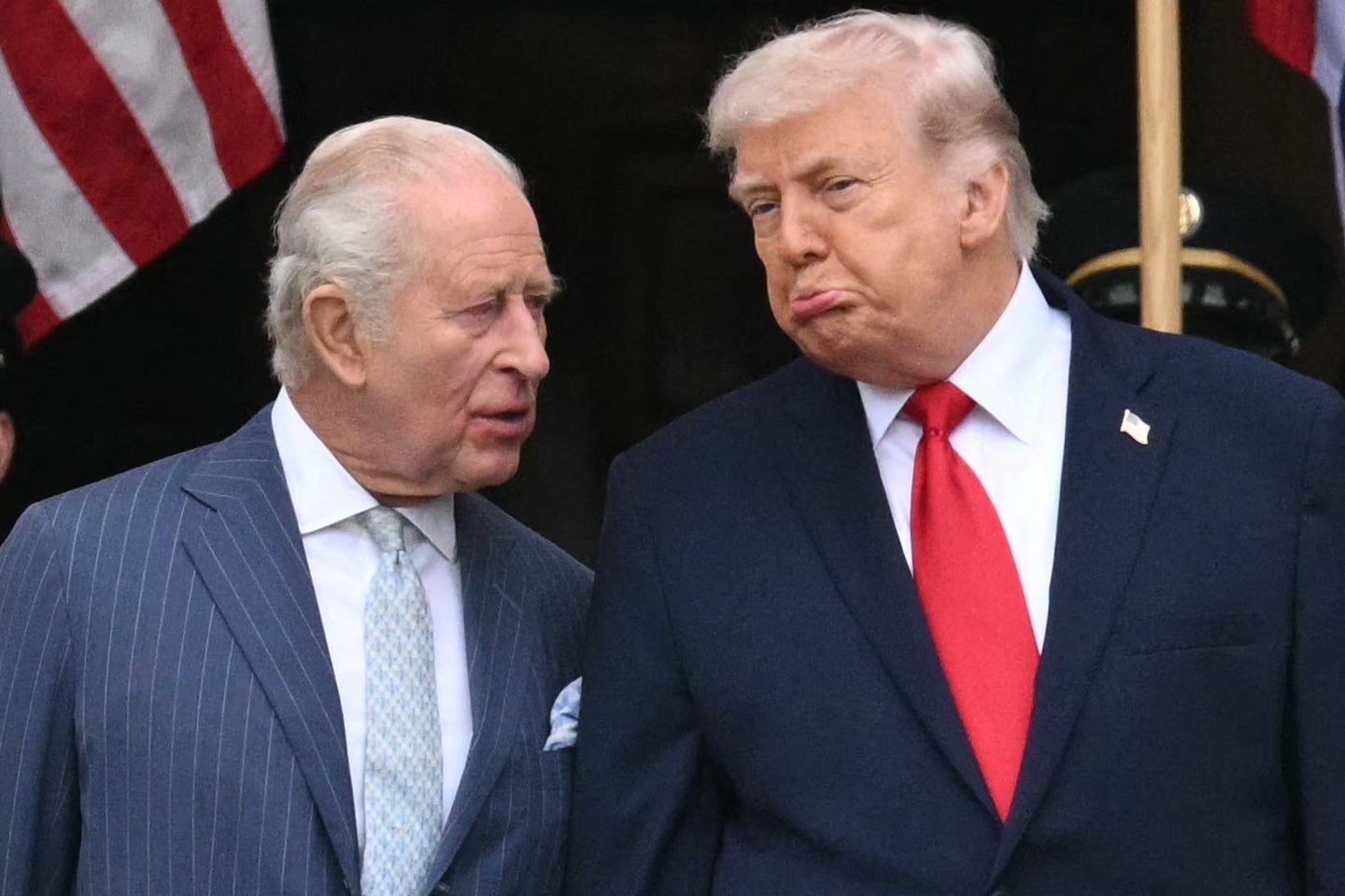 Kong Charles og Donald Trump er i dress og har en samtale; den ene har stripet dress, og den andre har rødt slips. De fremstår som engasjerte og uttrykksfulle; Trump rynker pannen med underleppen ute