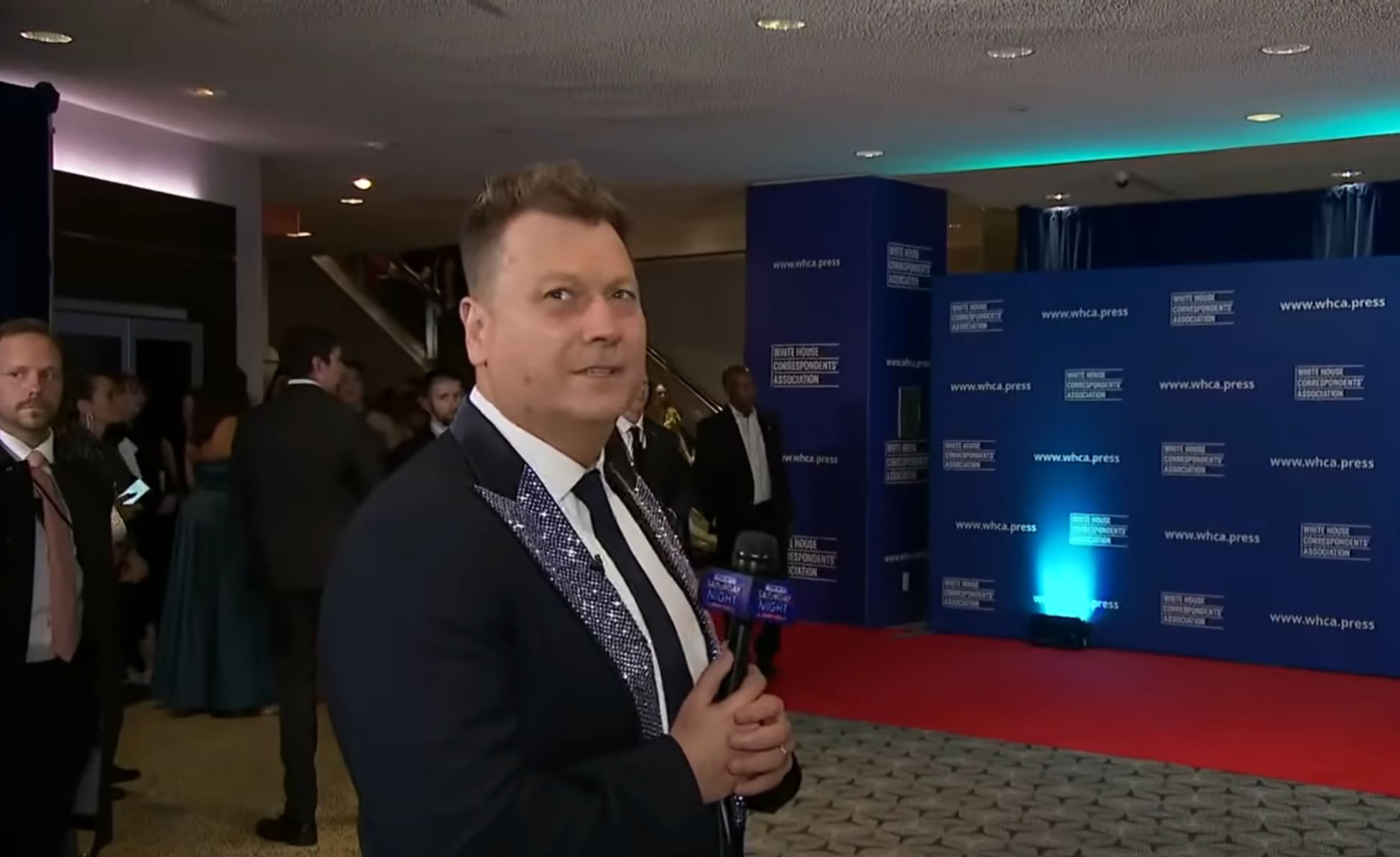 Person holding a microphone connected  a reddish  carpet astatine  an event, wearing a tuxedo with decorative lapels