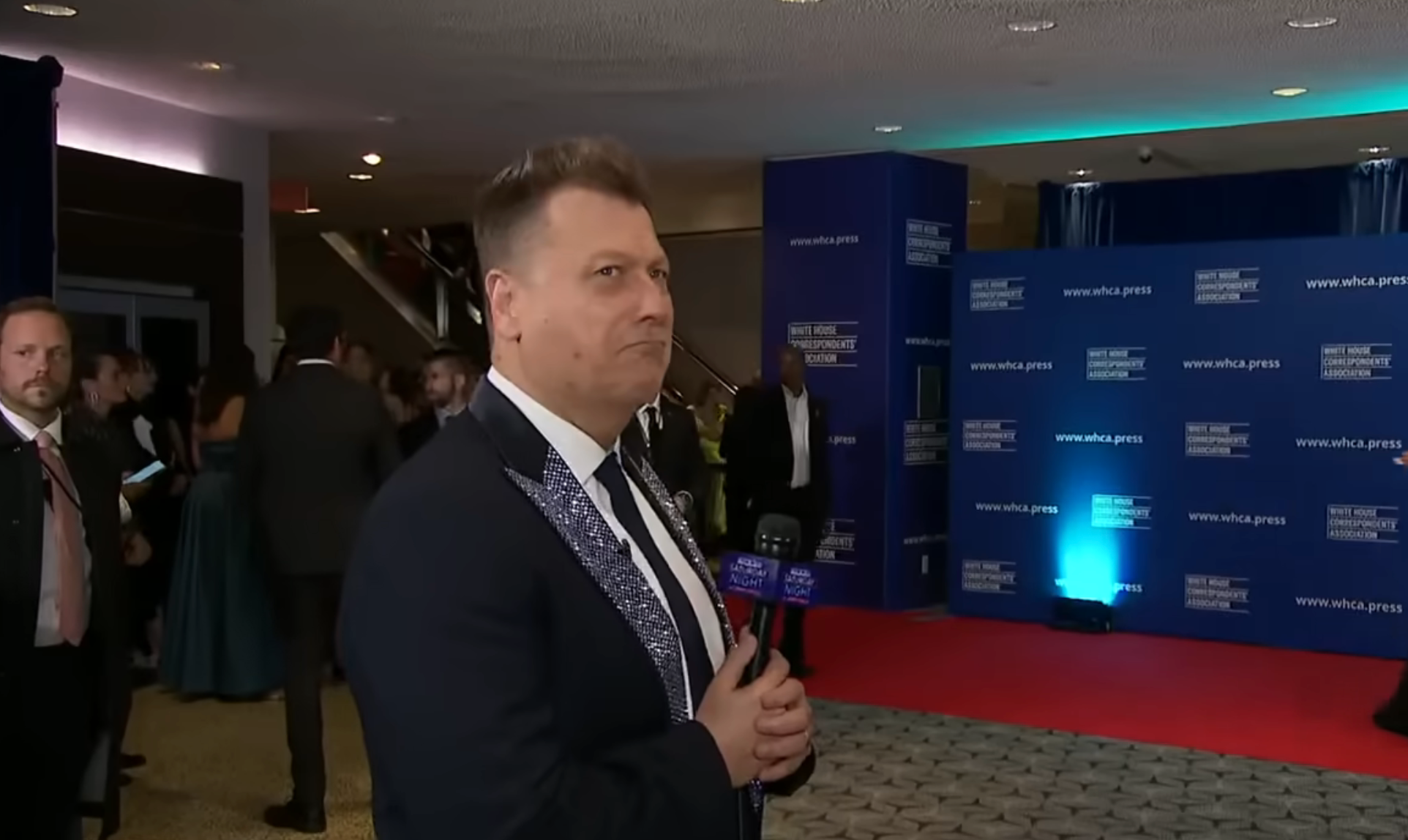 Person successful  a suit   with sequined lapels holding a microphone connected  a reddish  carpet astatine  a ceremonial  event