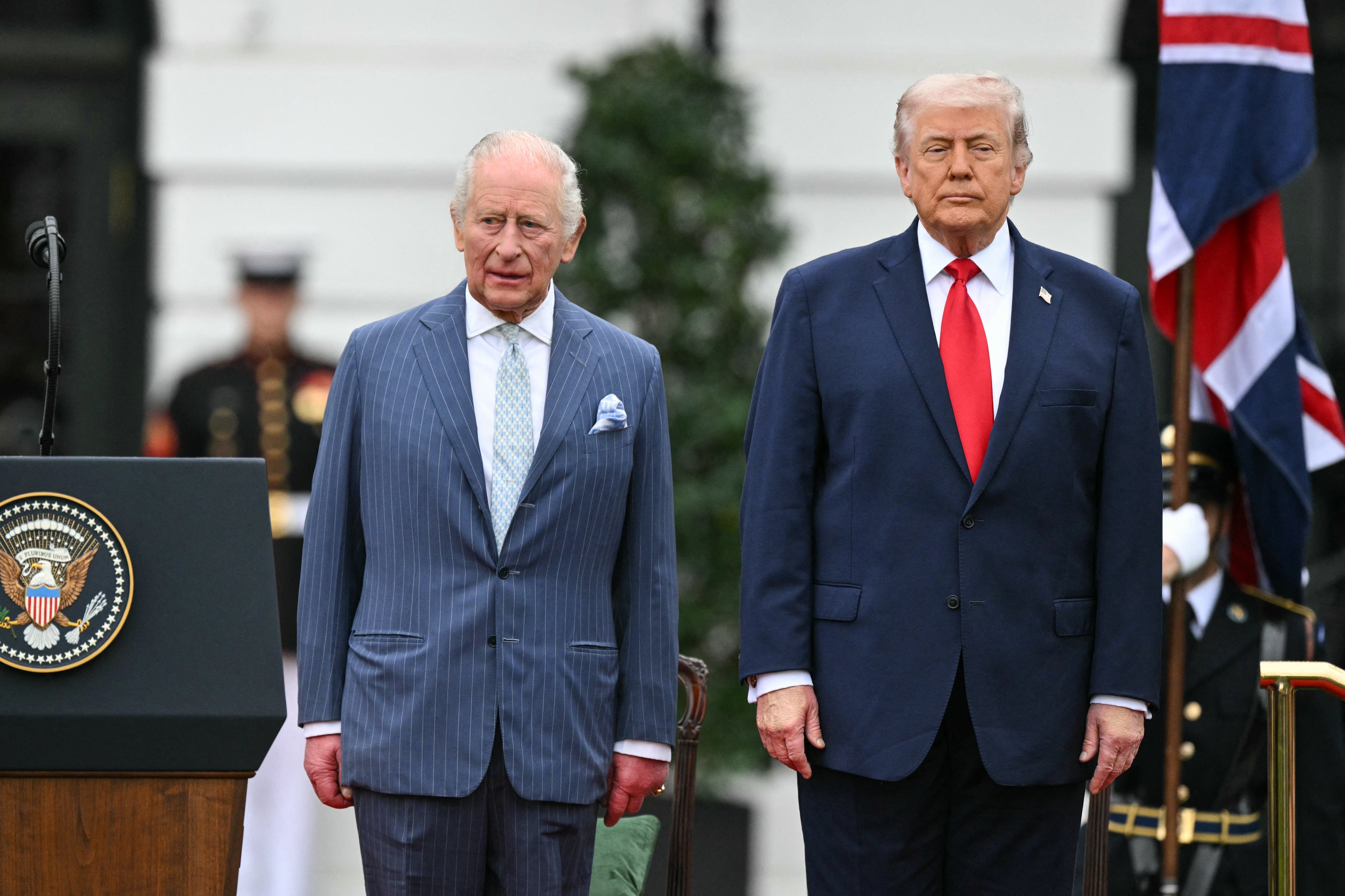 Kong Charles og Donald Trump i dress står side om side ved en formell begivenhet, med amerikanske og britiske flagg