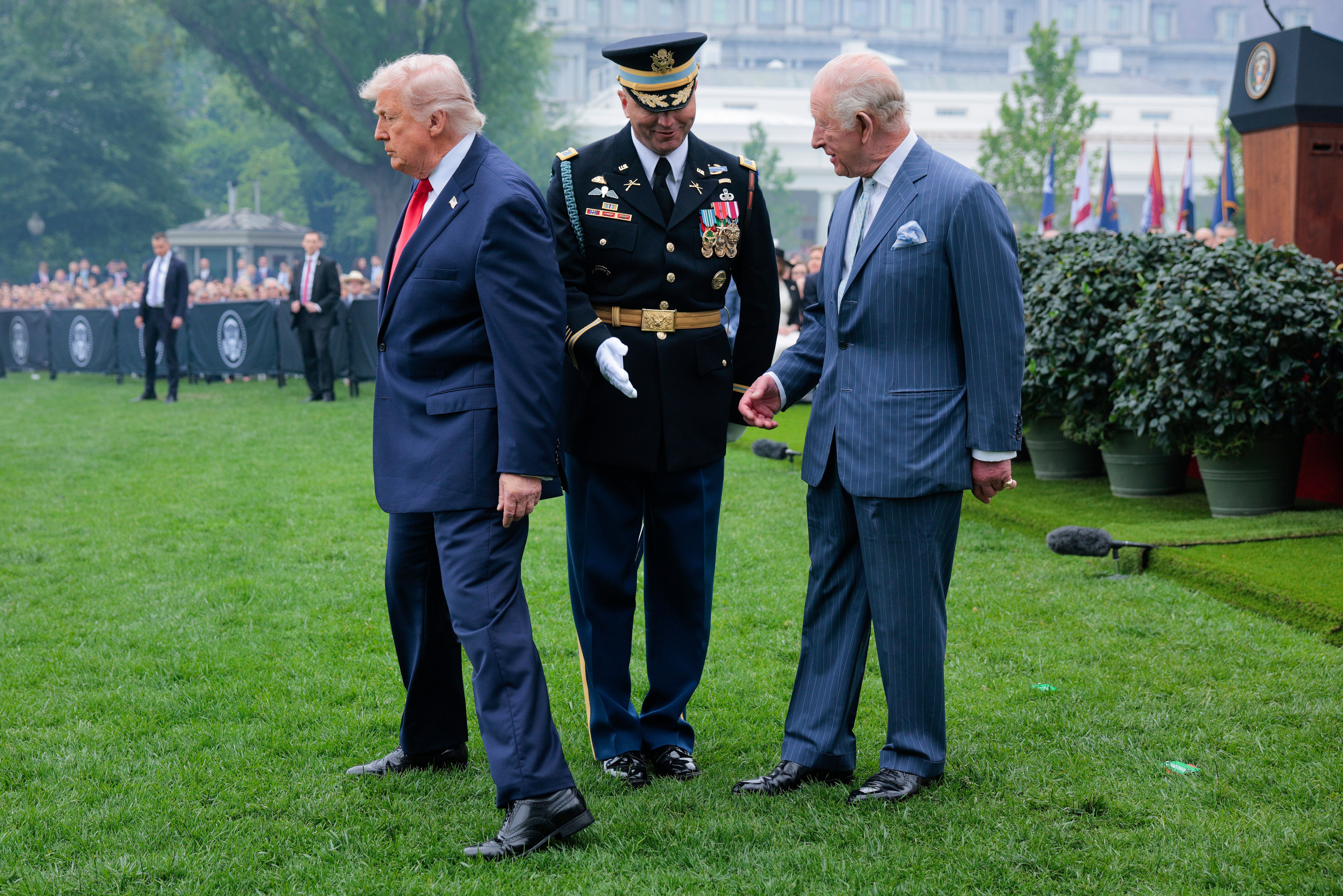 Donald Trump står keitete med bena spredt på plenen i Det hvite hus nær en uniformert marinesoldat og kong Charles; en folkemengde ser på