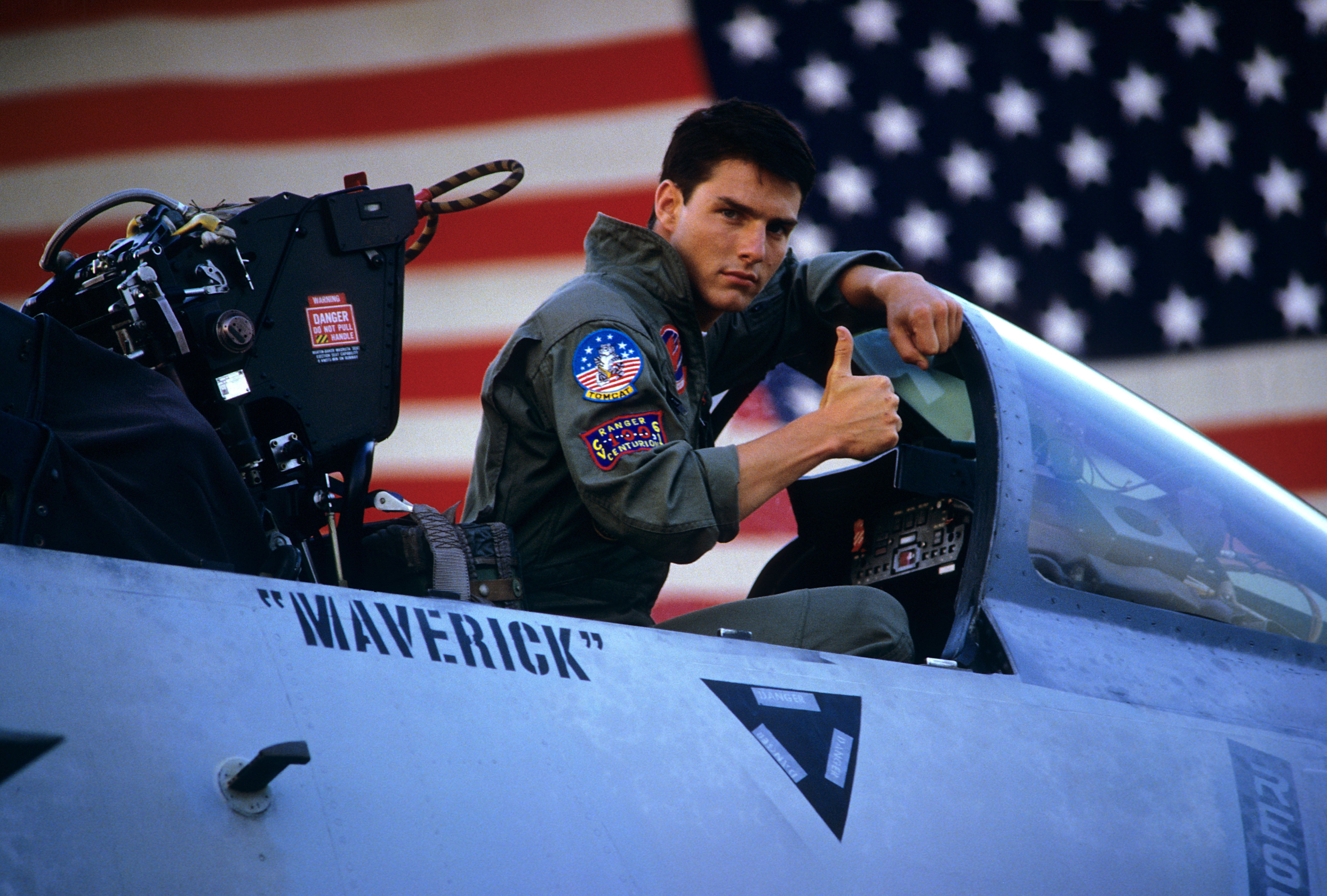 Person successful  a pilot's azygous   sits successful  a pitchy  cockpit, giving a thumbs up, with an American emblem  backdrop. The helmet reads "Maverick."