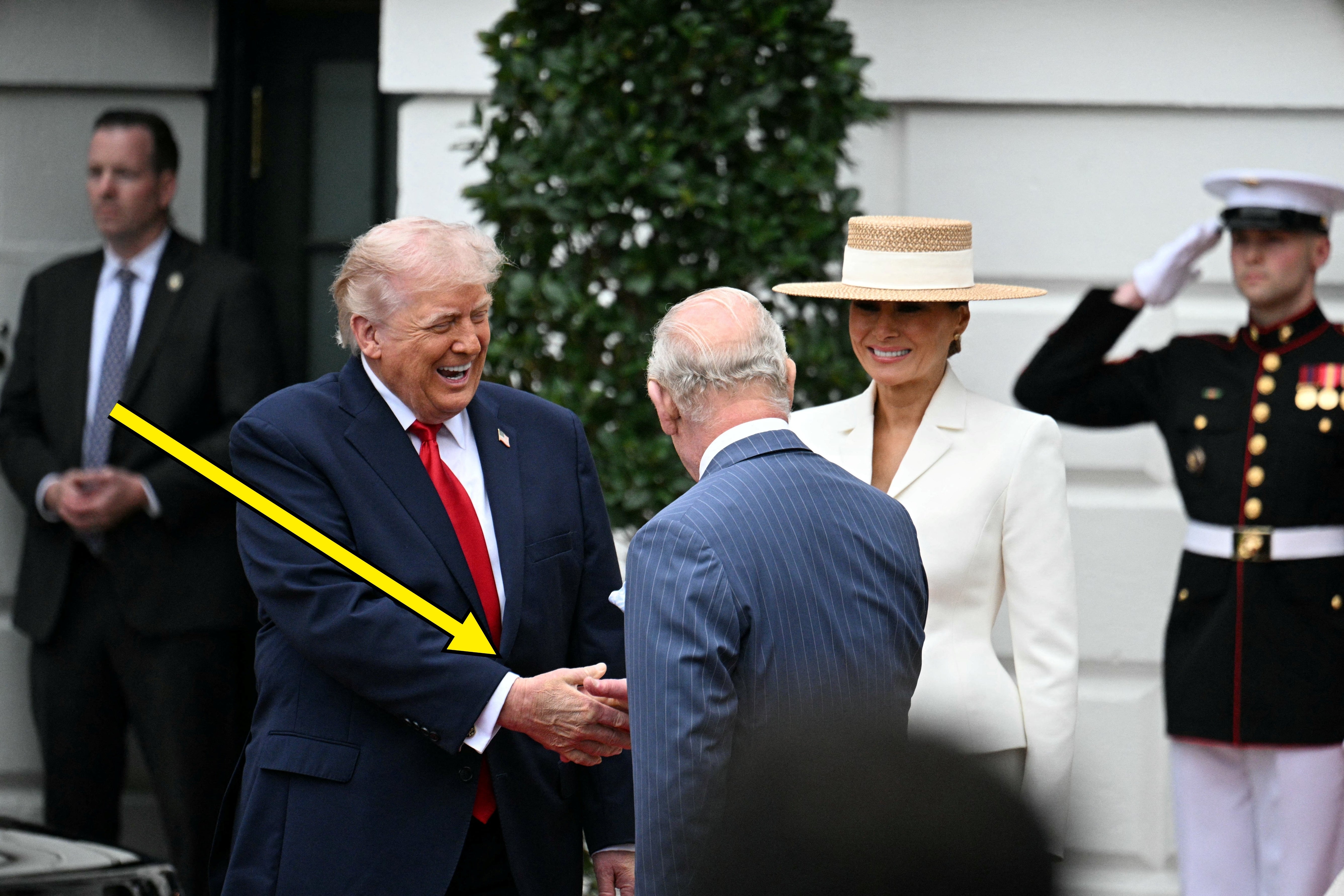 Donald Trump i dress håndhilser på kong Charles utendørs, med en Melania i hatt og en uniformert marinesoldat i nærheten
