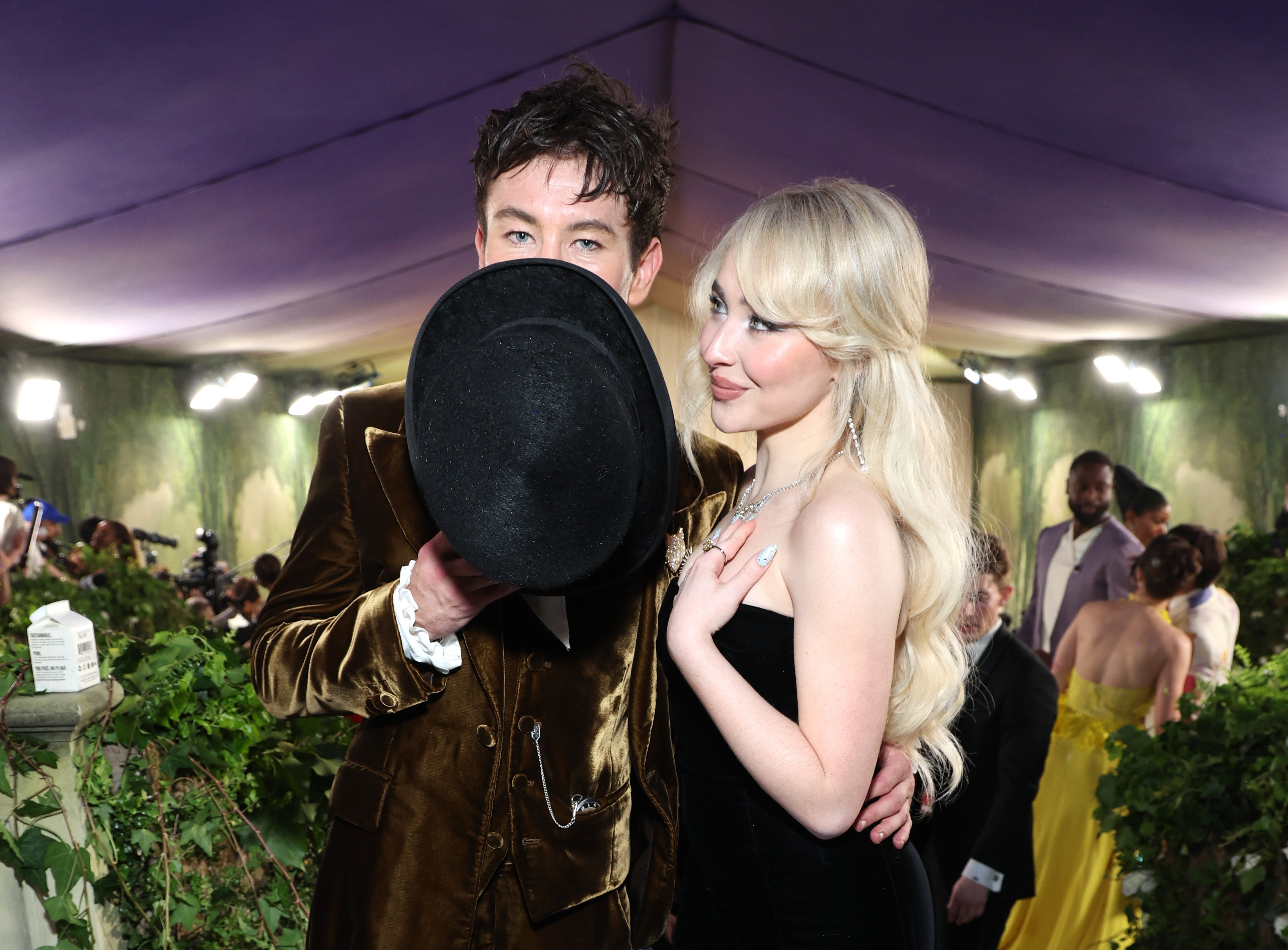 Two people at an event; one is holding a hat partially covering their face, while the other in a sleek dress smiles at the camera
