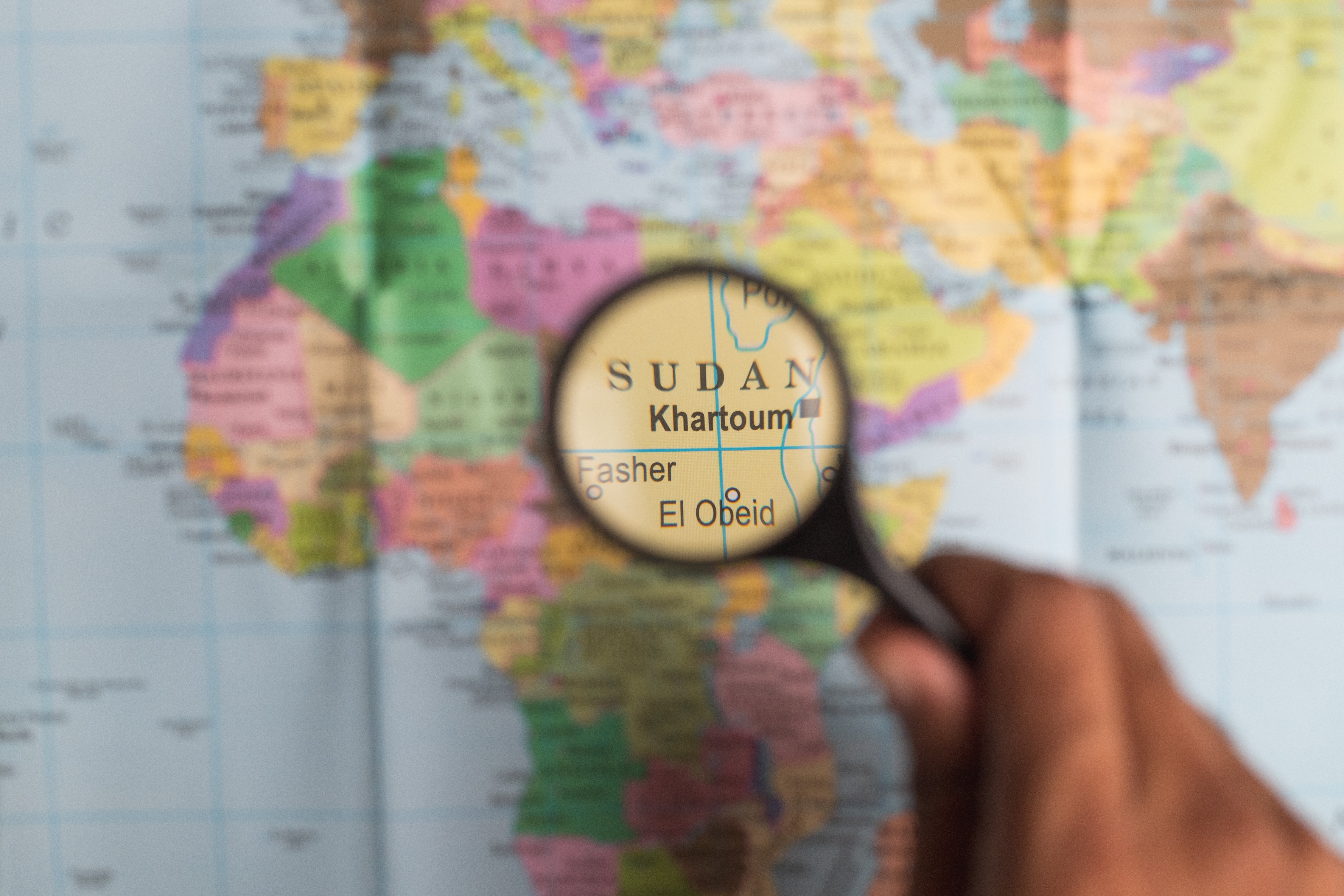 A hand holds a magnifying glass over a map, enlarging the area showing Sudan and its capital, Khartoum