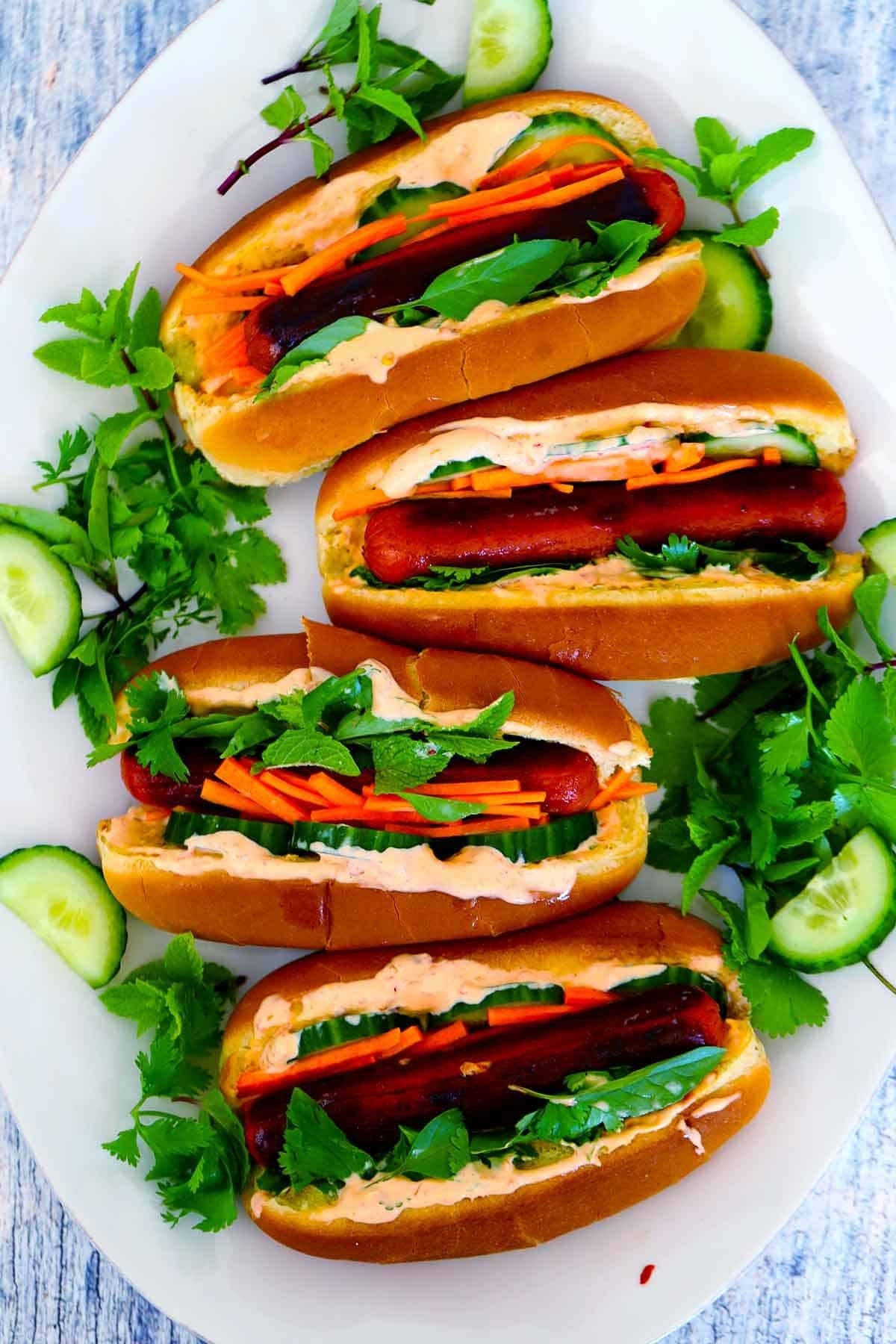 Four gourmet hot dogs in buns, topped with sliced cucumbers, carrots, and cilantro, served on a white platter