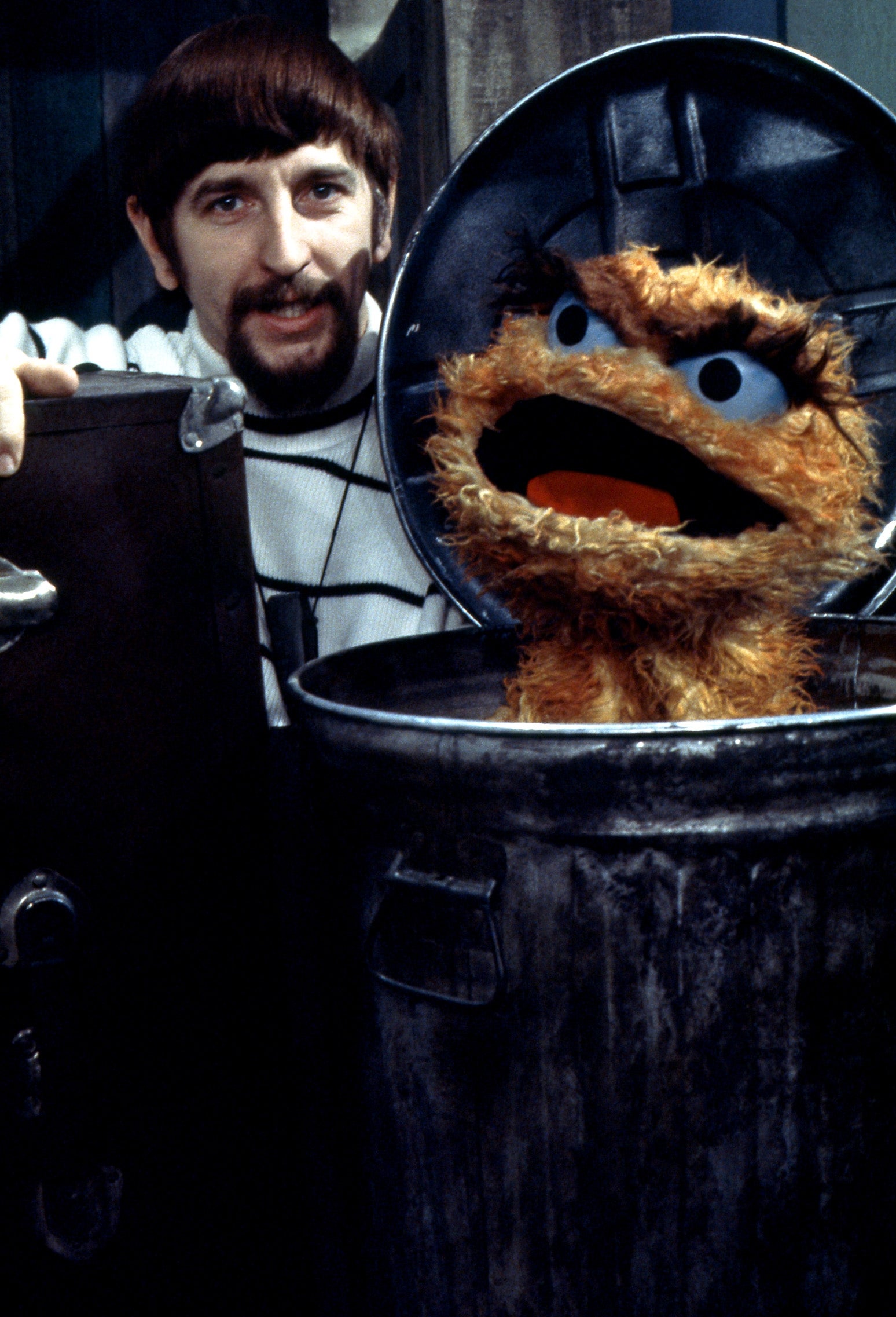 A antheral   with facial hairsbreadth  smiles arsenic  helium  opens a trash tin  containing a fuzzy puppet with bushy eyebrows, apt  from a retro TV show