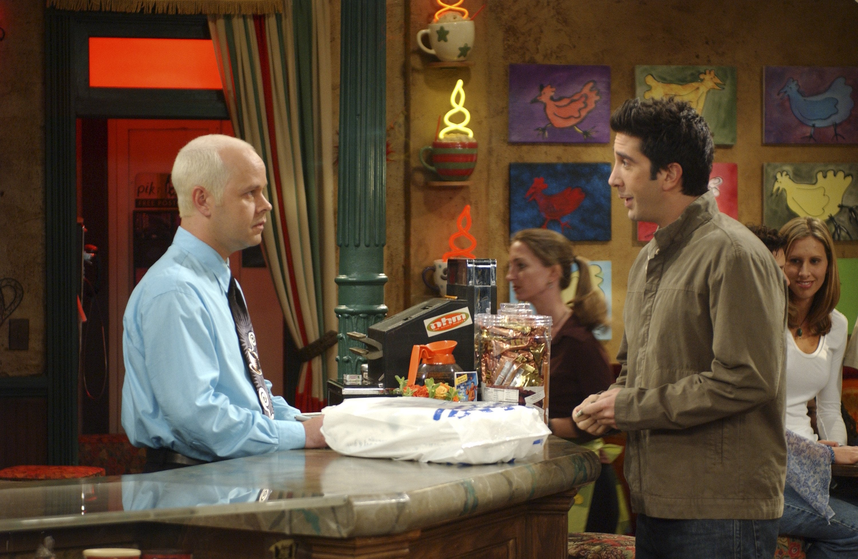 Two men are talking astatine  a java  store  counter. One is wearing a bluish  garment  and tie, the different   a casual jacket. Background shows customers seated