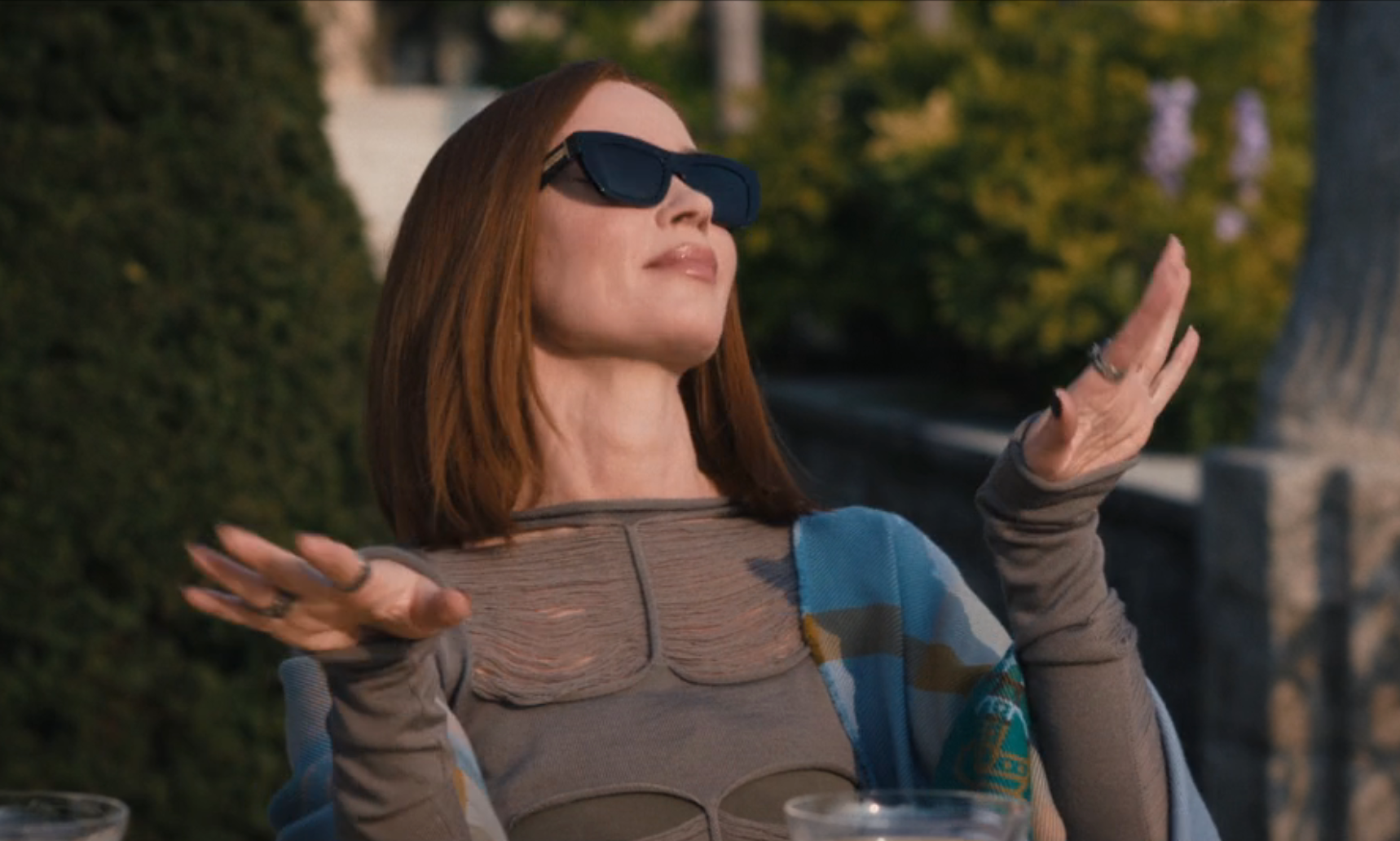 A idiosyncratic   successful  fashionable sunglasses and a stylish layered outfit poses confidently, arms open, astatine  an outdoor gathering