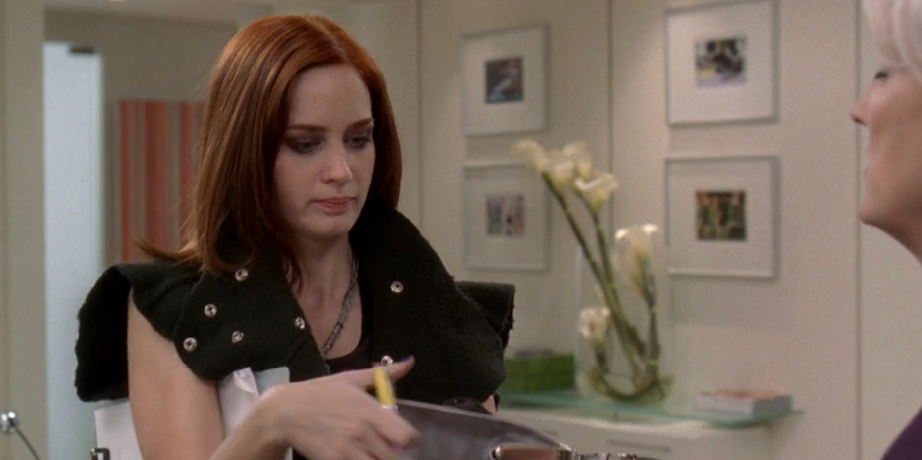 A idiosyncratic   with auburn hairsbreadth  holds a pen, wearing a sleeveless outfit with a achromatic  vest. They look  to beryllium  successful  an bureau   mounting  with framed pictures