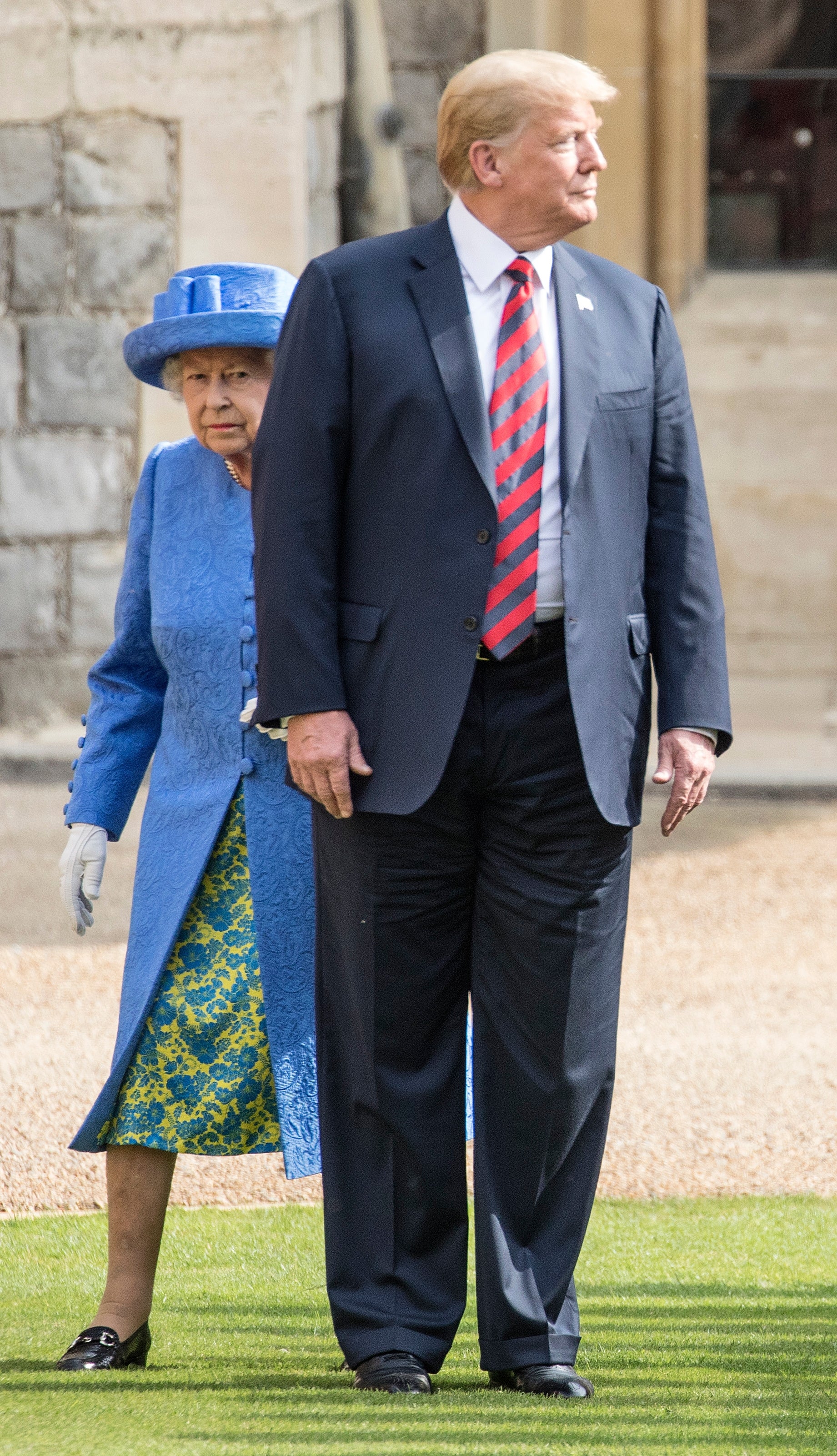En kvinne i mønstret antrekk og hatt går med en mann i dress og stripet slips på en gresssti utenfor en historisk bygning