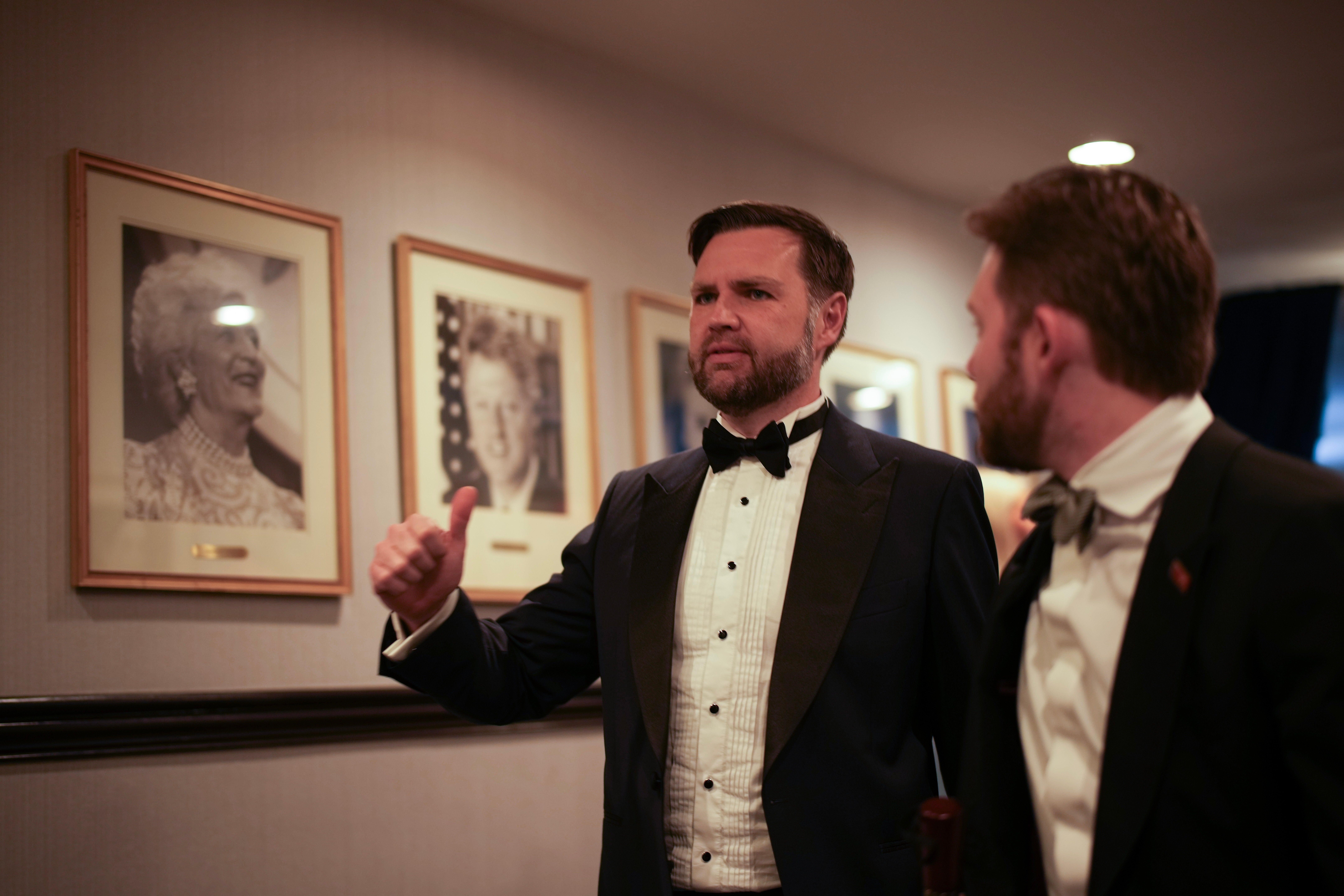 To menn i smoking står i et galleri med innrammede portretter på veggen. Man gestikulerer med tommel opp