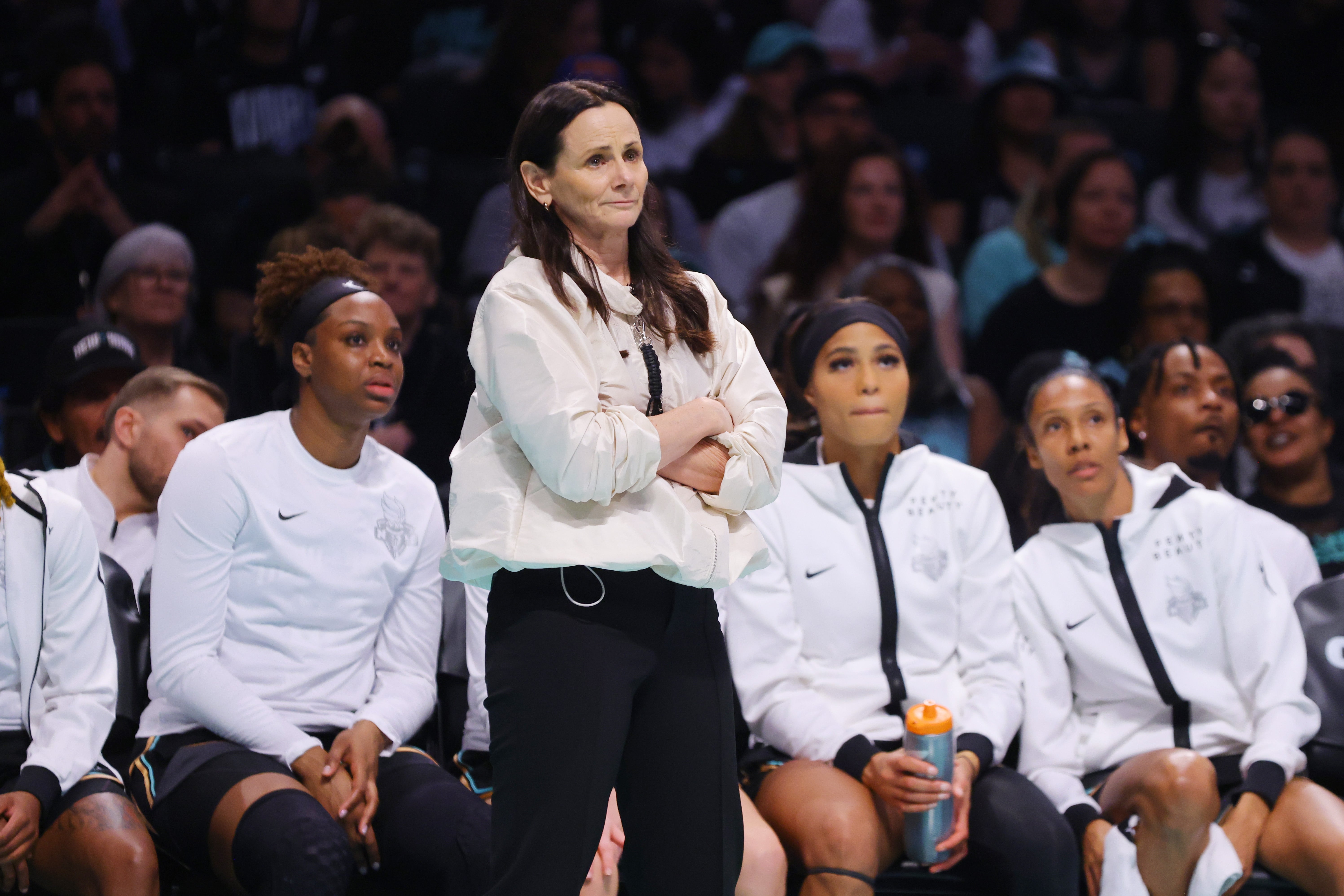 A hoops  manager  successful  a achromatic  jacket, arms crossed, stands successful  beforehand   of seated players during a game. Players deterioration  achromatic  jackets and achromatic  shorts